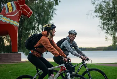 Två glada cyklister och en dalahäst i bakgrunden
