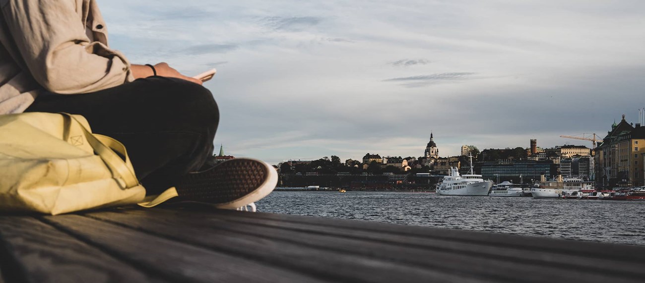 En person sitter på en brygga och håller en mobil med utsikt över Stockholm