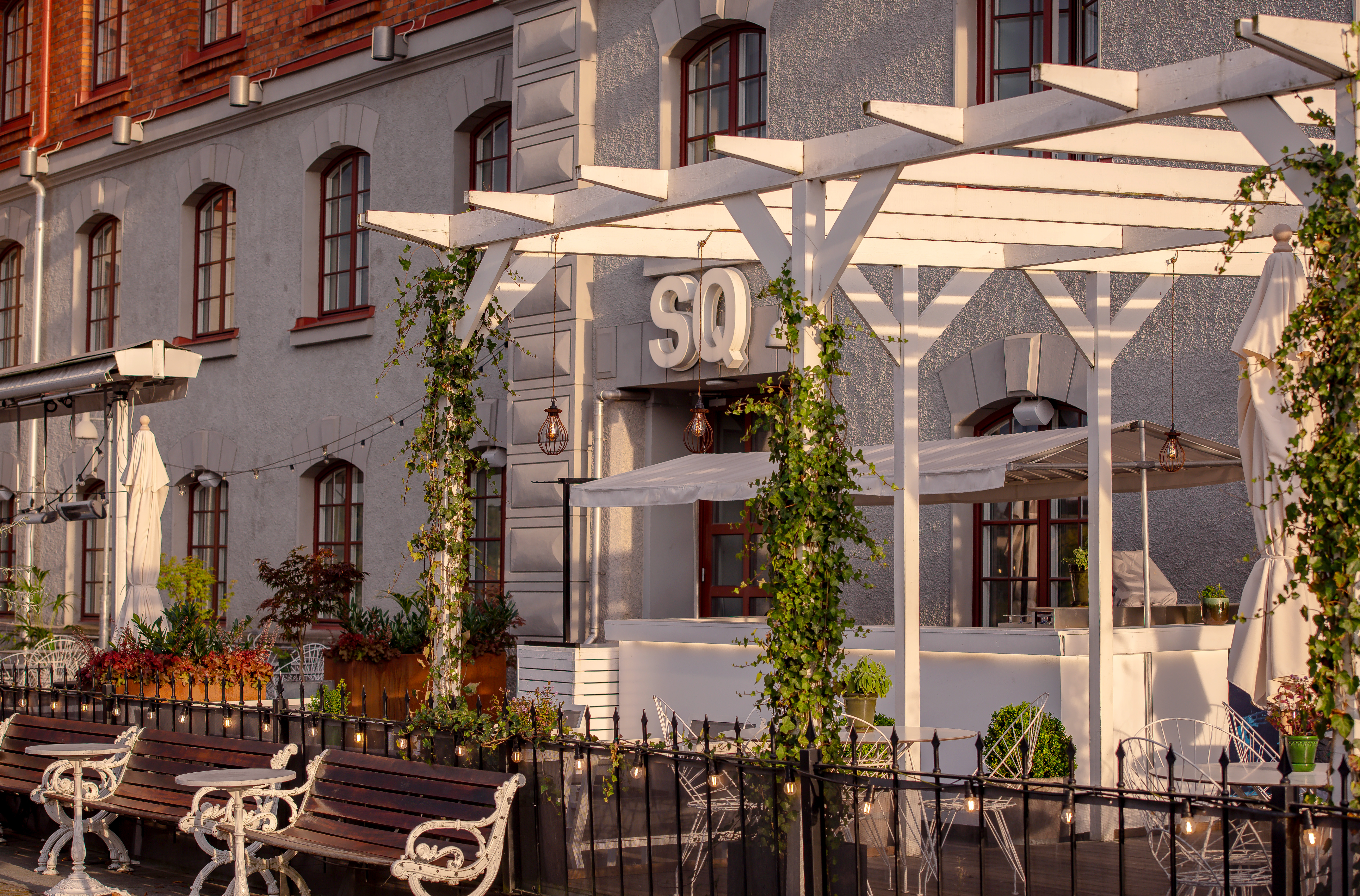 Exteriör av en charmig restaurang med vit pergola och gröna växter, omgiven av träbänkar och historisk byggnad i bakgrunden under solnedgång