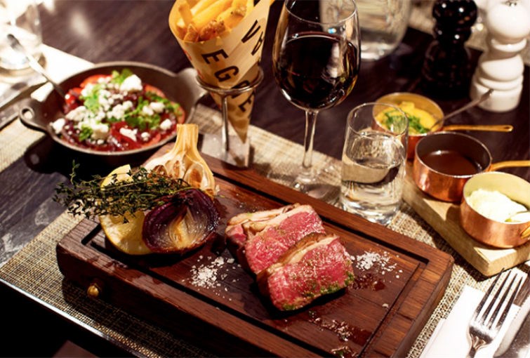 Köttbitar serverad på planka och ett vinglas med rött vin bredvid