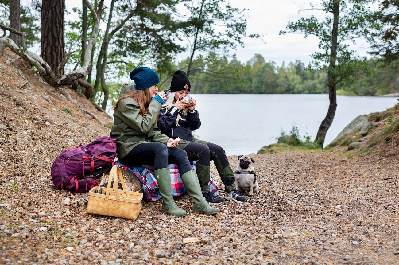 Två kvinnor och en liten hund har en fikapaus i en skogsdunge vid vattnet