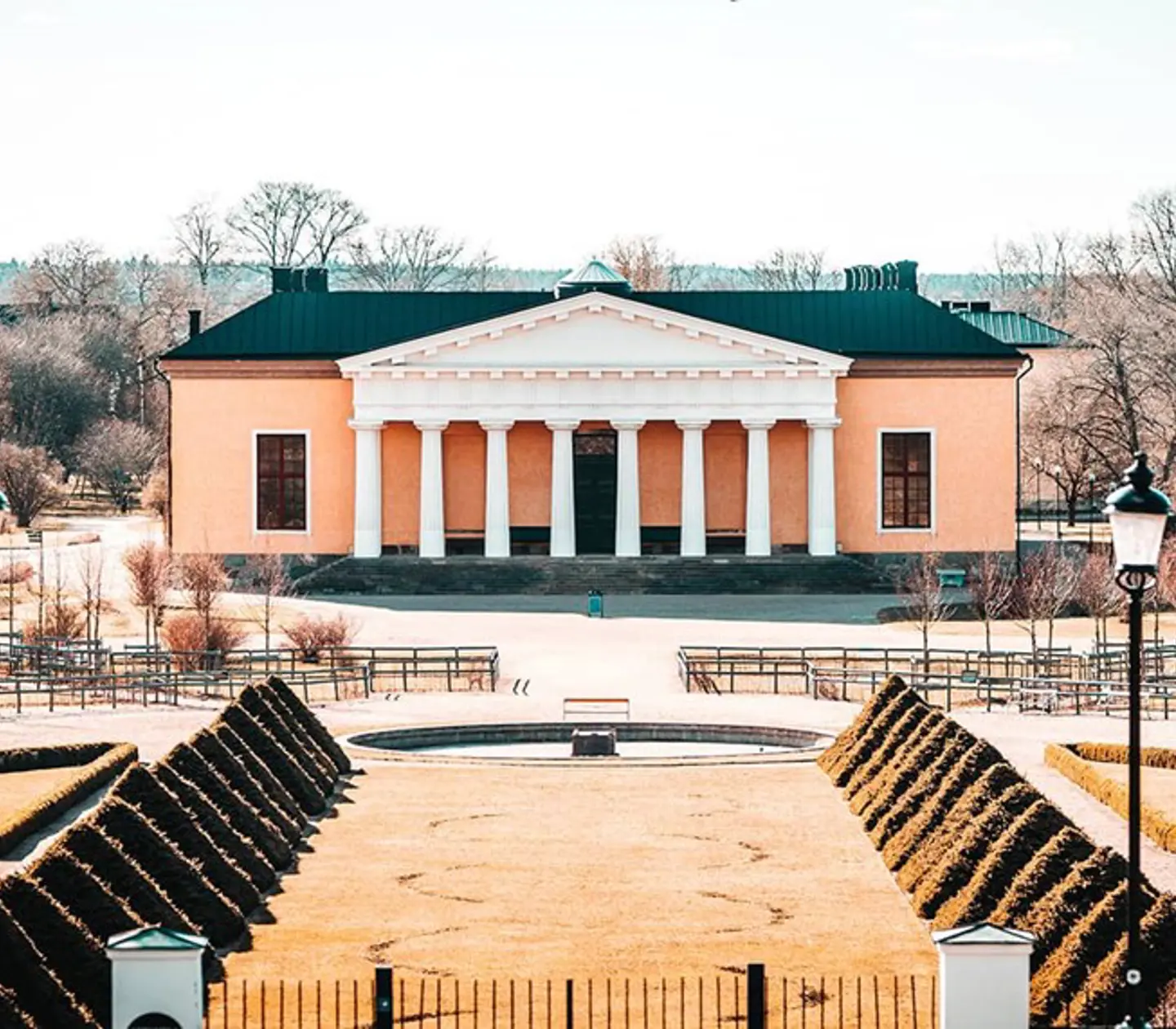 Hus med putsad, rosa fasad och vita pelare, omgiven av en stor trädgård med buskar och träd
