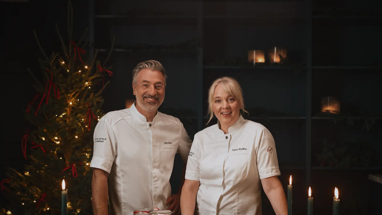Tareq Taylor & Carina Brydling in white uniforms stand smiling at a festive holiday table adorned with candles, greenery, and a decorated Christmas tree in the background.