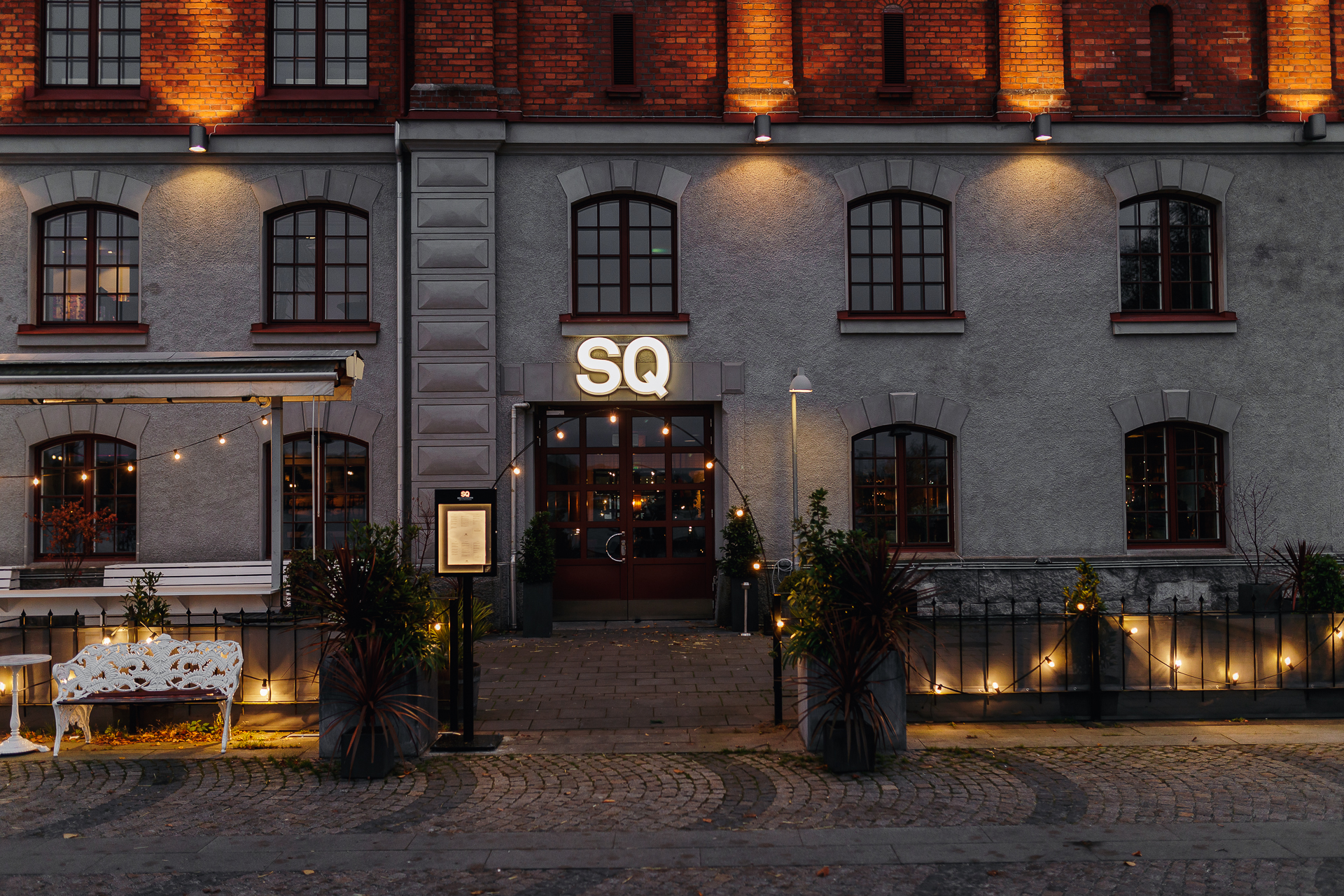 Historisk byggnad med restaurangentré i kvällsbelysning, röd tegelvägg och fönster med välvda detaljer, modern uteservering med dekorativ belysning.