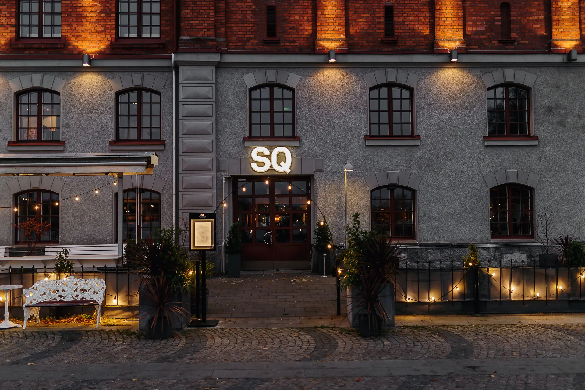 Historic building with string lights decorating the entrance and a sign reading "SQ" above the door. A white bench and potted plants enhance the cozy atmosphere.