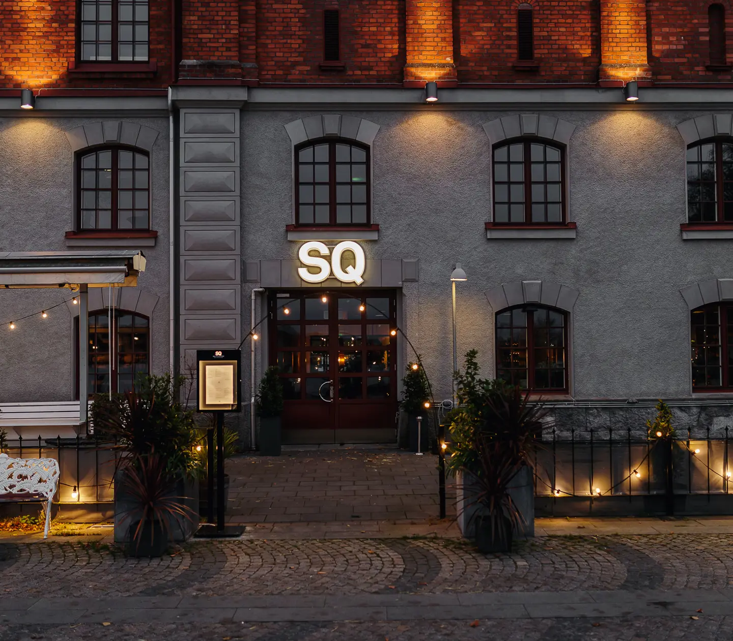 Historisk byggnad med restaurangentré i kvällsbelysning, röd tegelvägg och fönster med välvda detaljer, modern uteservering med dekorativ belysning.