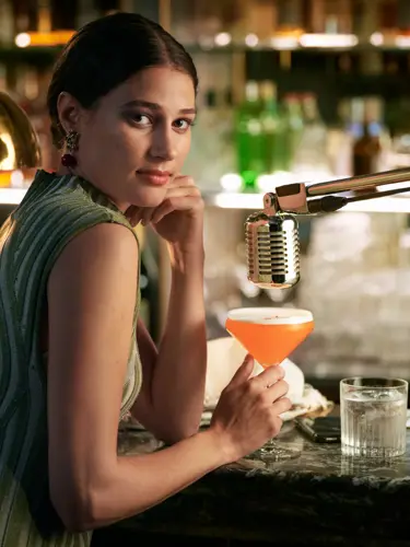 Woman in green dress sitting at bar with orange drink in hand and microphone over