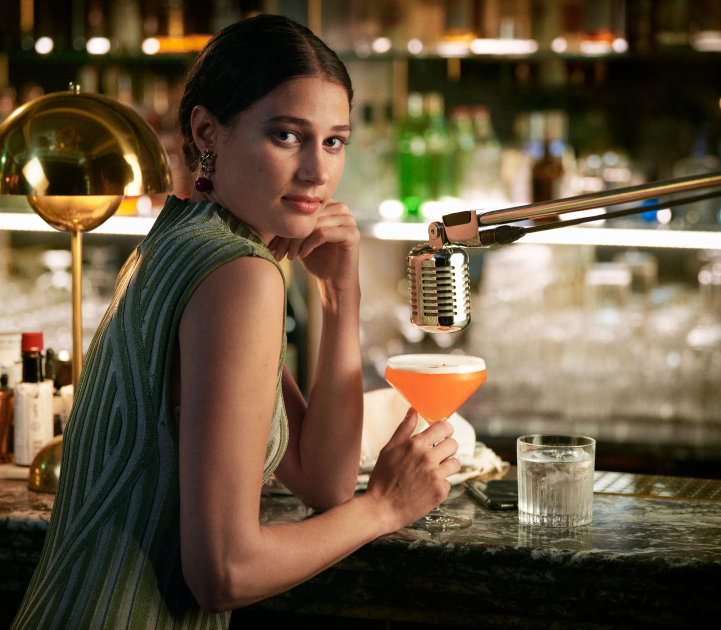Woman in green dress sitting in bar with orange drink in hand and microphone above