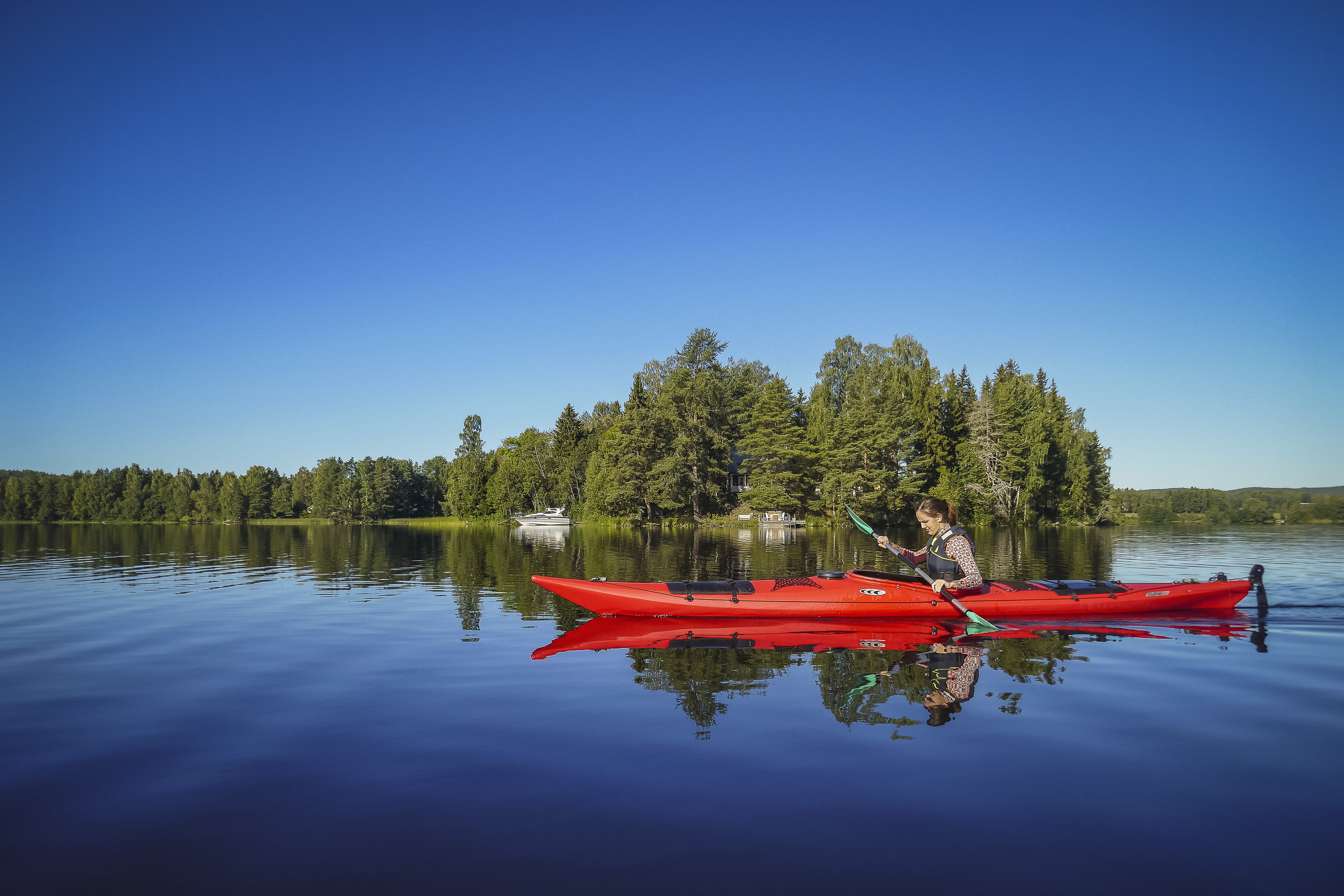 A person paddles a red kayak on a calm lake in front of a lush forest under a clear blue sky in Sweden
