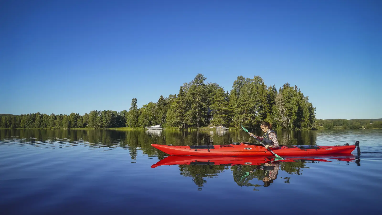 Person paddlar röd kajak på lugn sjö framför grönskande skog under klarblå himmel i Sverige