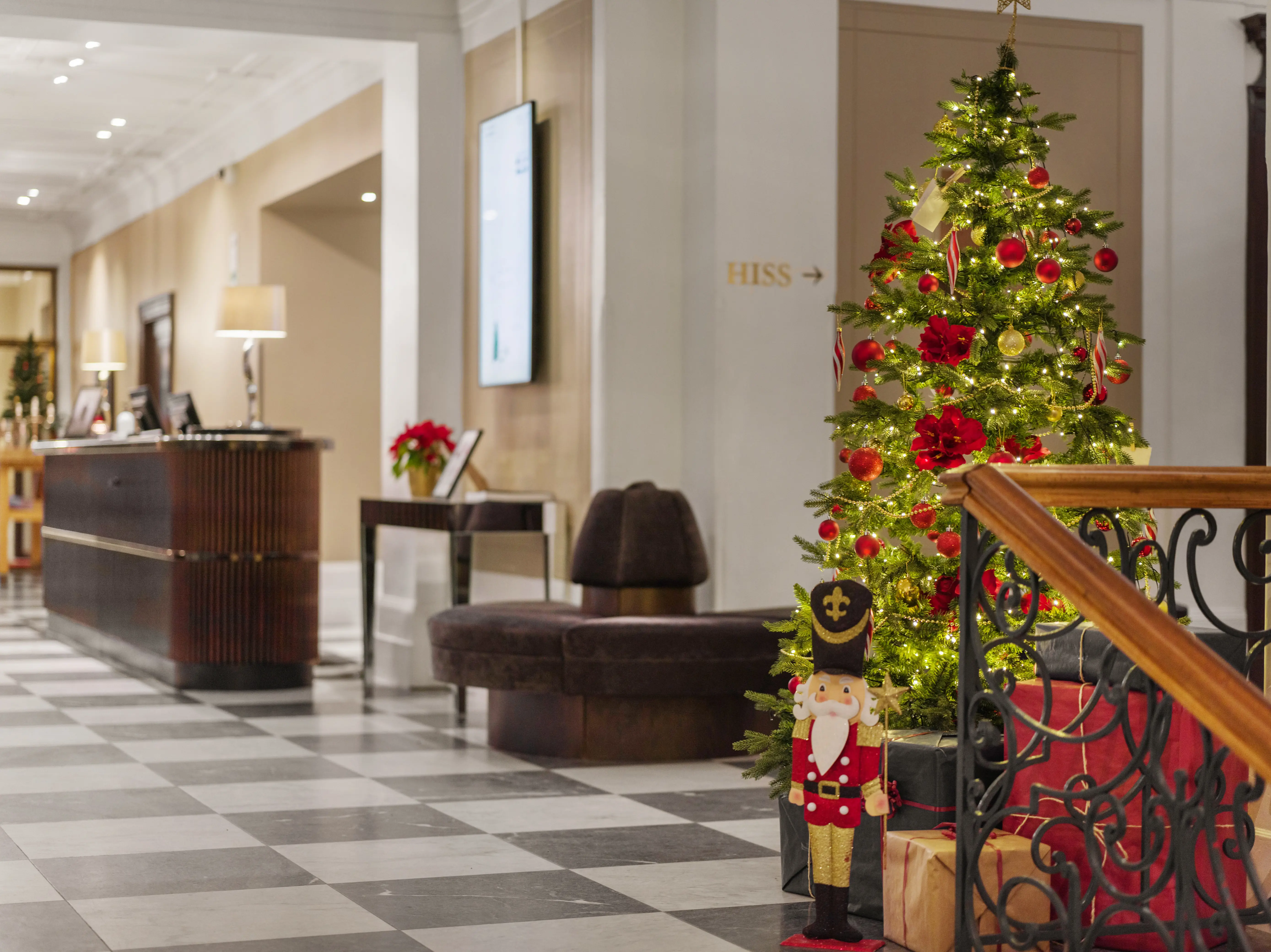 Julegran dekorerad med röda och guldiga kulor i hotellobby, bredvid en reception och en leksakssoldat.