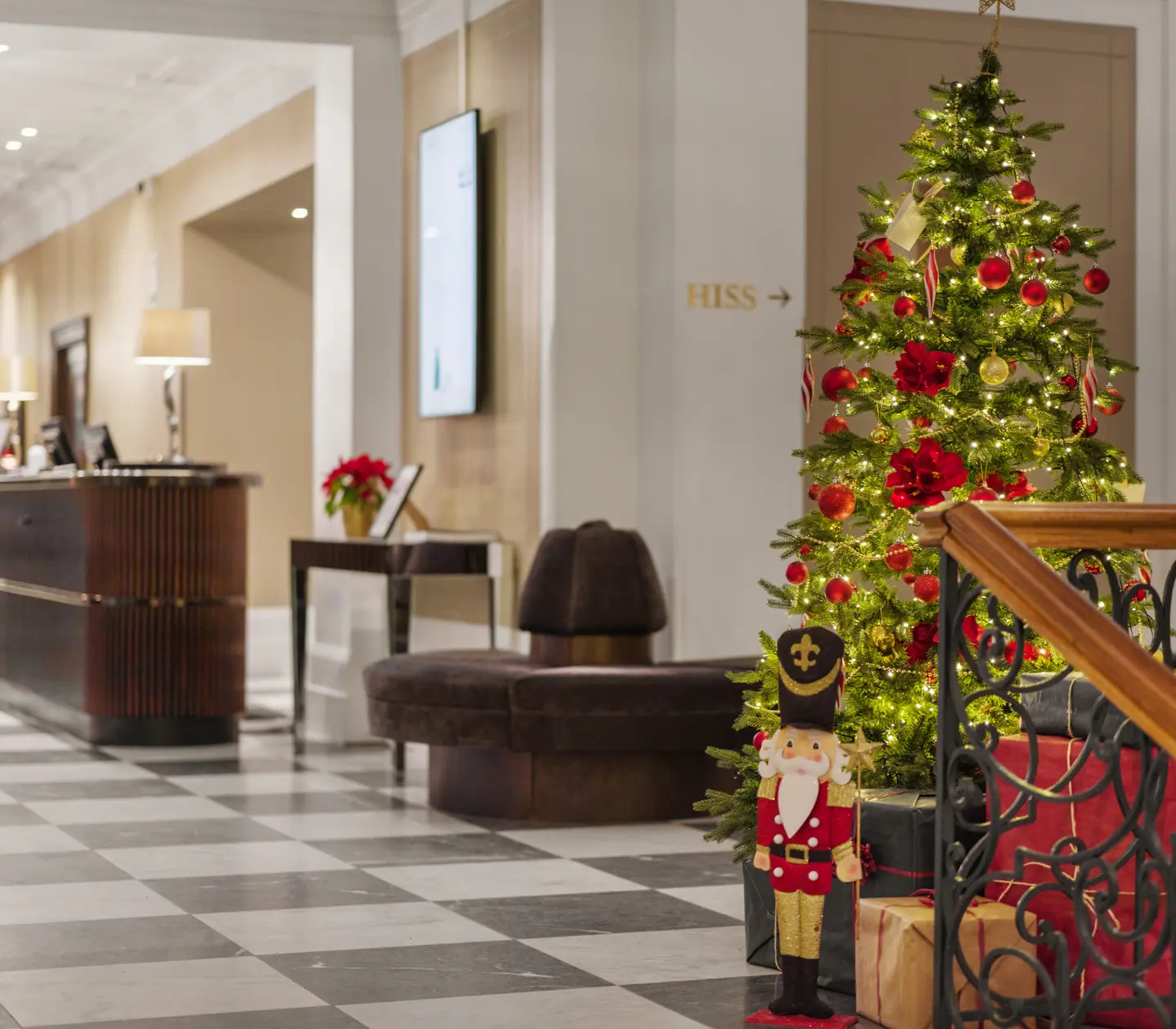 Julegran dekorerad med röda och guldiga kulor i hotellobby, bredvid en reception och en leksakssoldat.