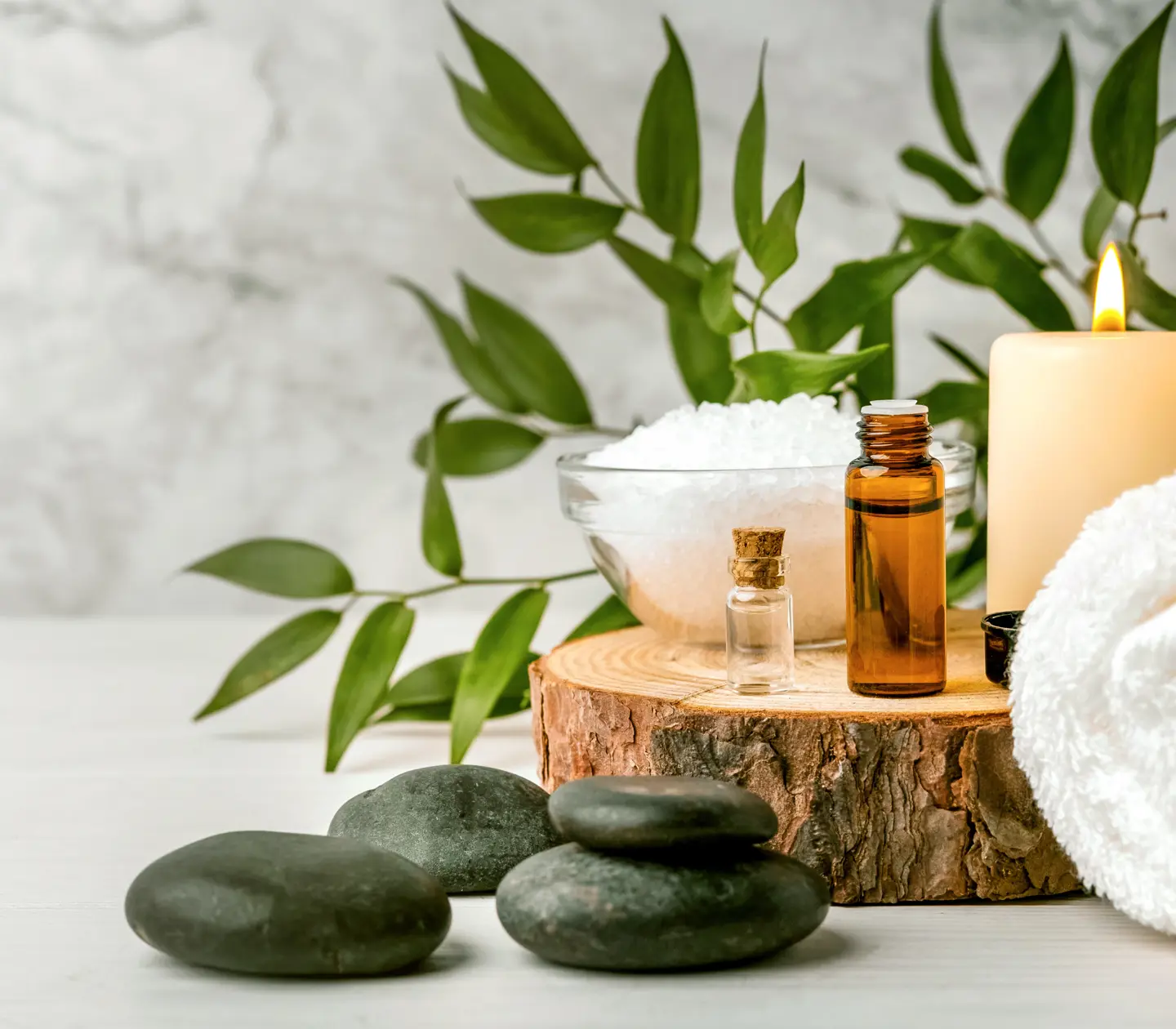 Spa essentials with candles, essential oils, bath salt, towel, and green leaves on wooden surface against marble background.