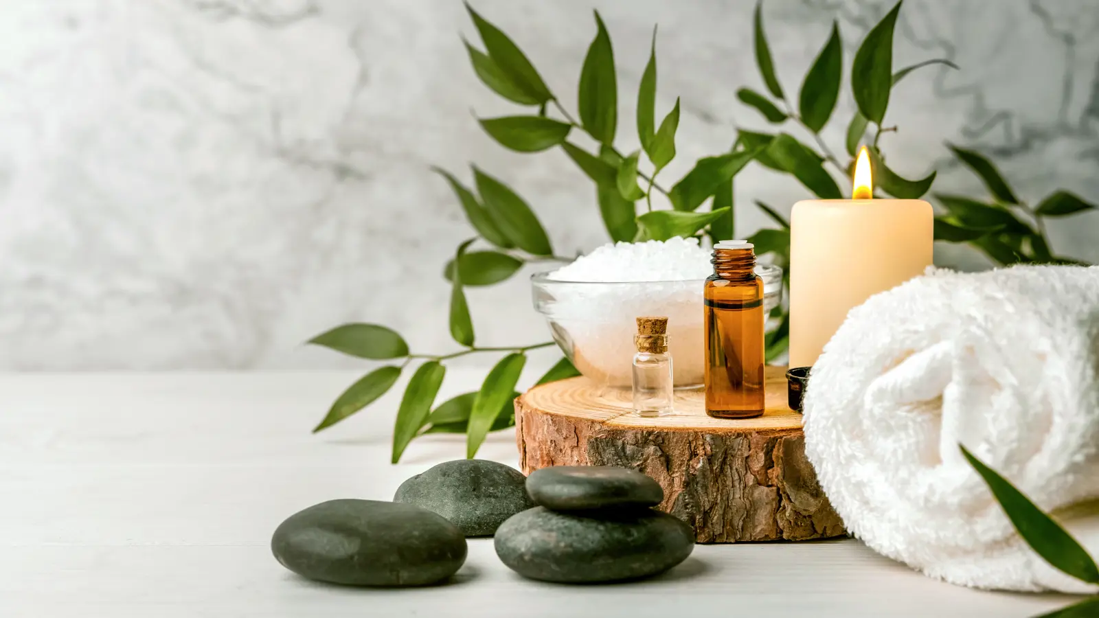 Spa essentials with candles, essential oils, bath salt, towel, and green leaves on wooden surface against marble background.