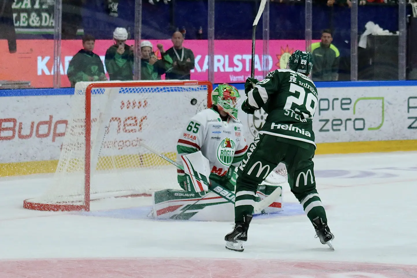 Hockey player in green and white jersey attempting to score against a goalie in a competitive ice hockey match, with cheering fans in the background