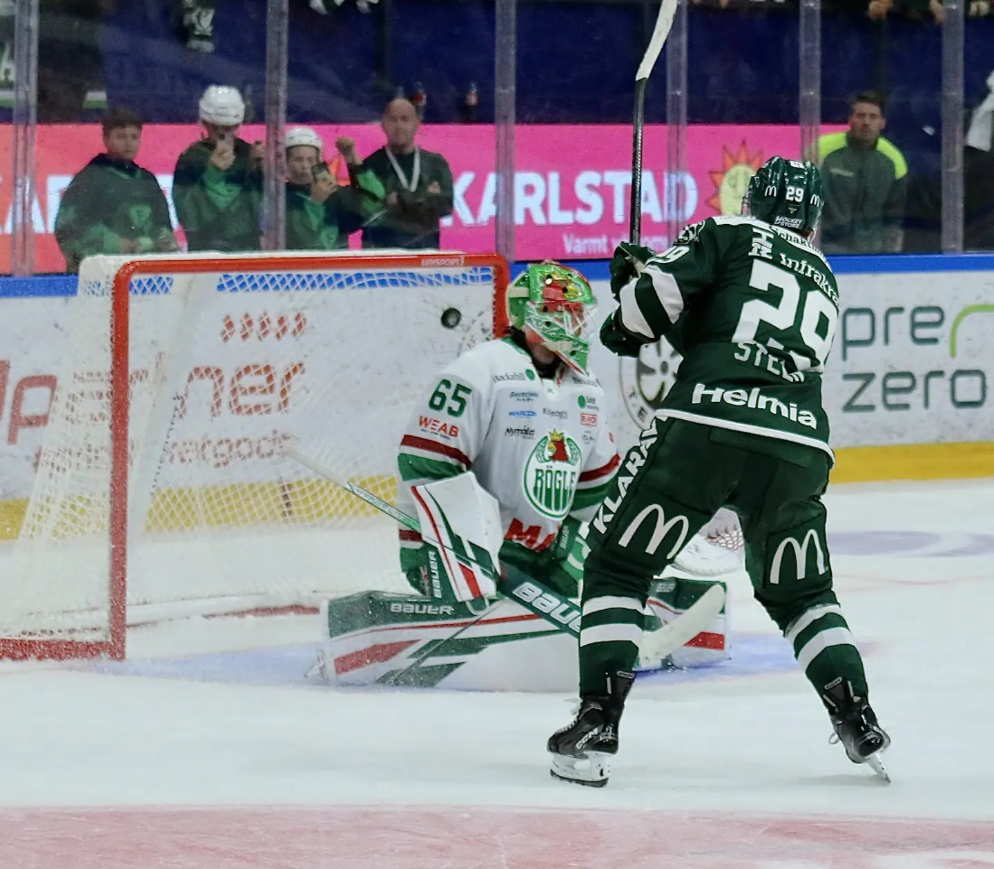 Hockey player in green and white jersey attempting to score against a goalie in a competitive ice hockey match, with cheering fans in the background