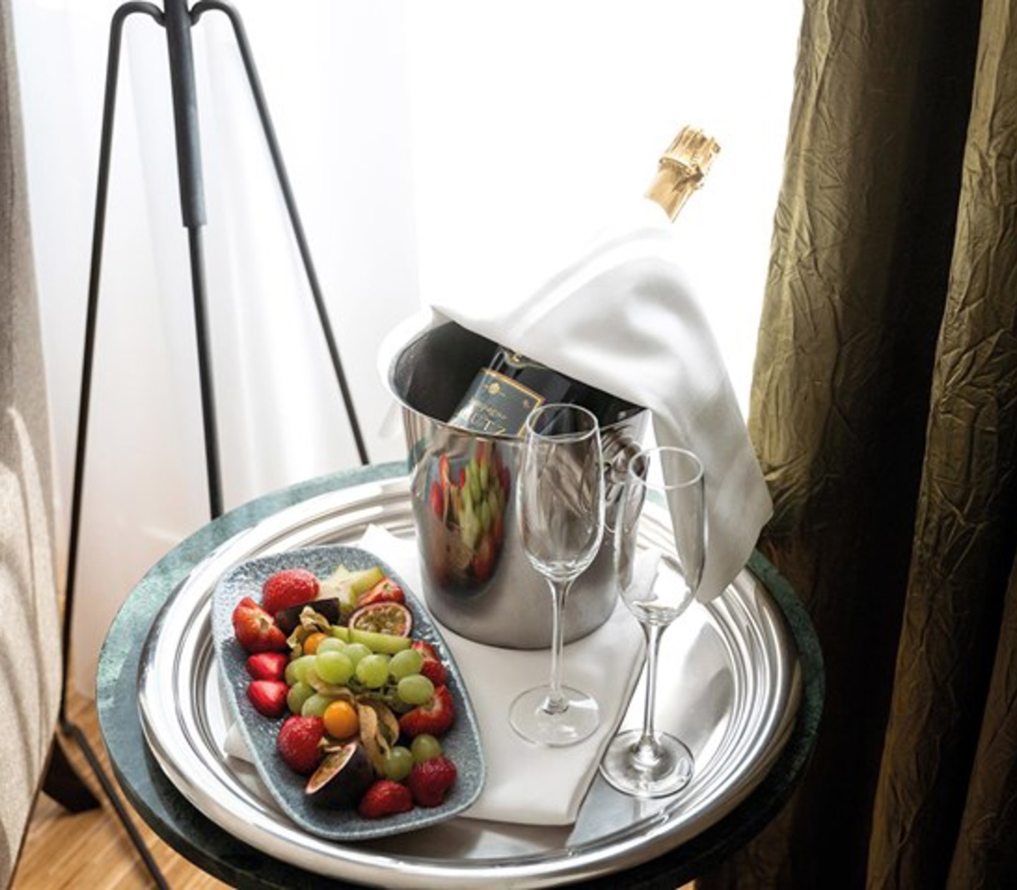 A bottle of Champagne in an ice bucket, two champagne glasses and a fruit platter on a table