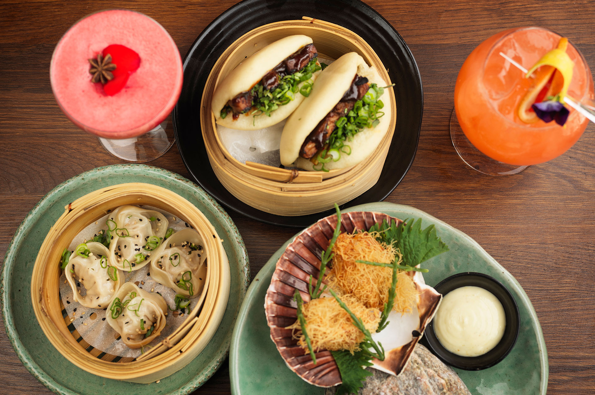 Asian dishes with bao buns, dumplings, and fried seafood on a table, served with two colorful cocktails