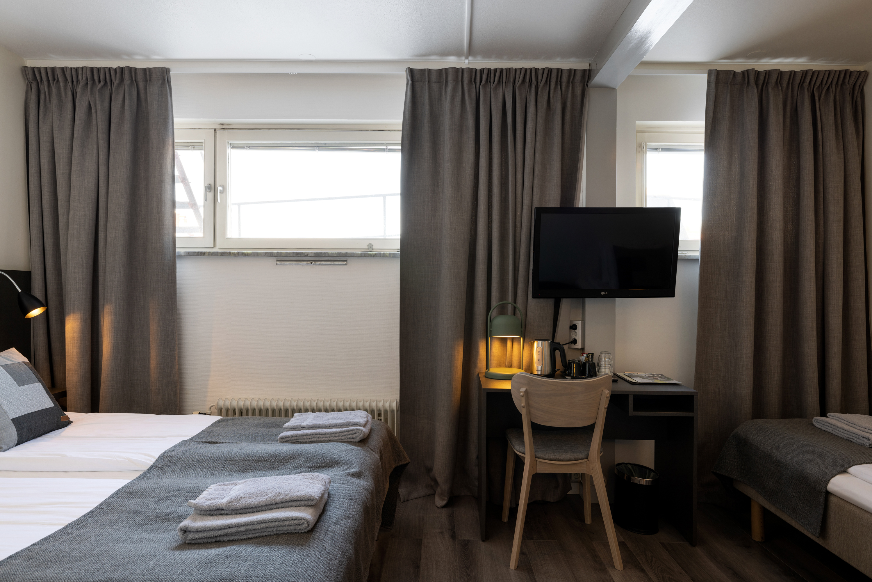 Ljust hotellrum med två enkelsängar med sänggavel i trä, tv, fönster och gardiner