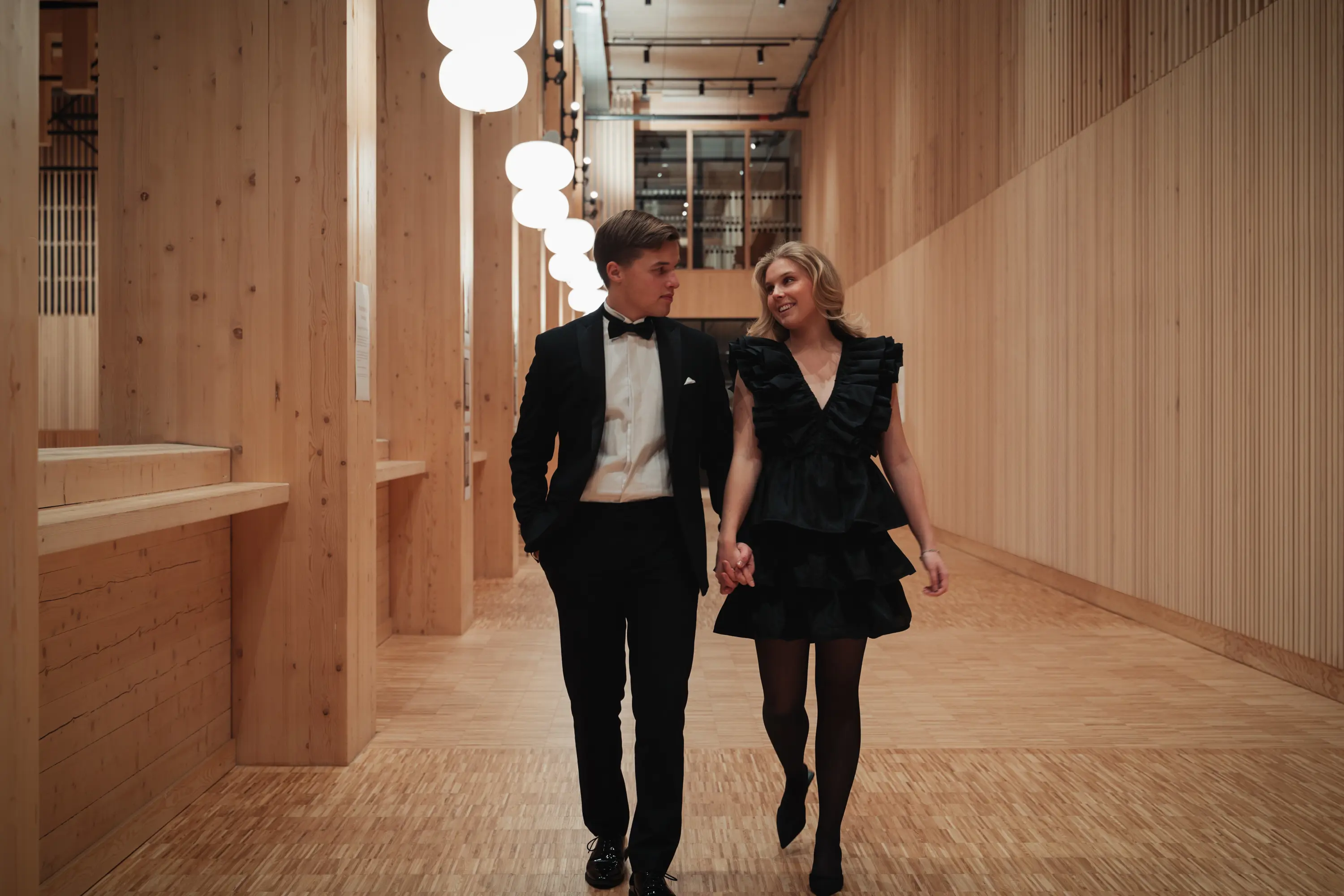 Ett par i formell klädsel, en man i frack och en kvinna i svart klänning, promenerar hand i hand genom en elegant träkorridor.