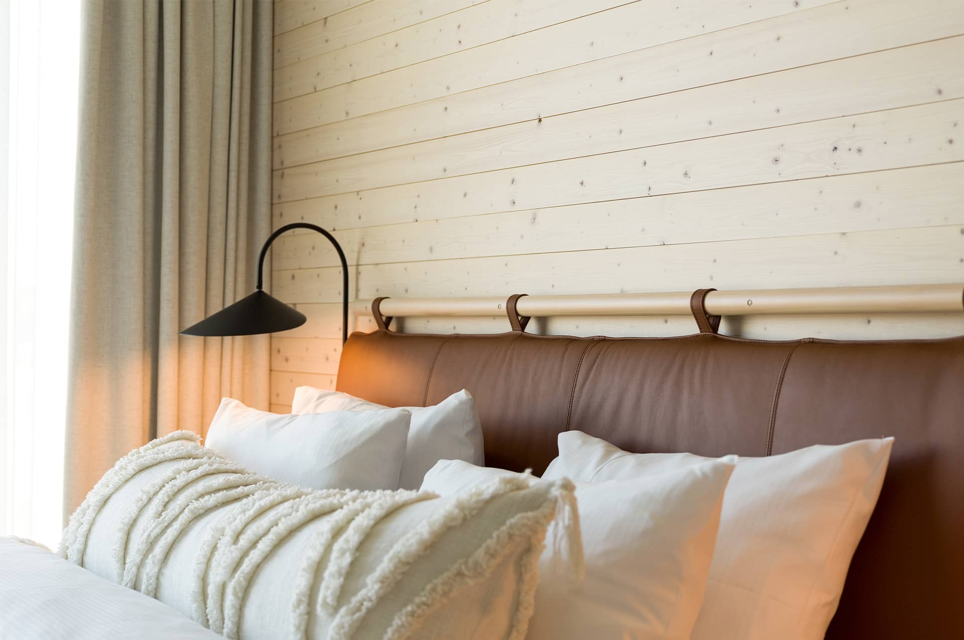 Fluffigt bäddad säng med sänggavel i brunt läder som står framför en vägg med ljus träpanel