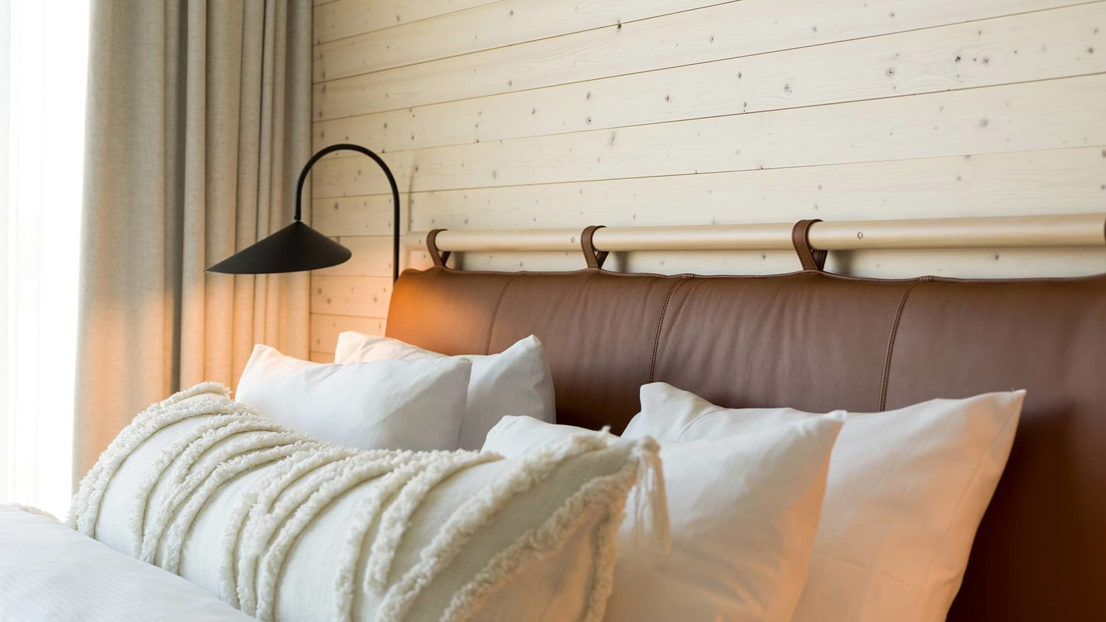 Fluffy bed with brown leather headboard standing in front of a light wood paneled wall