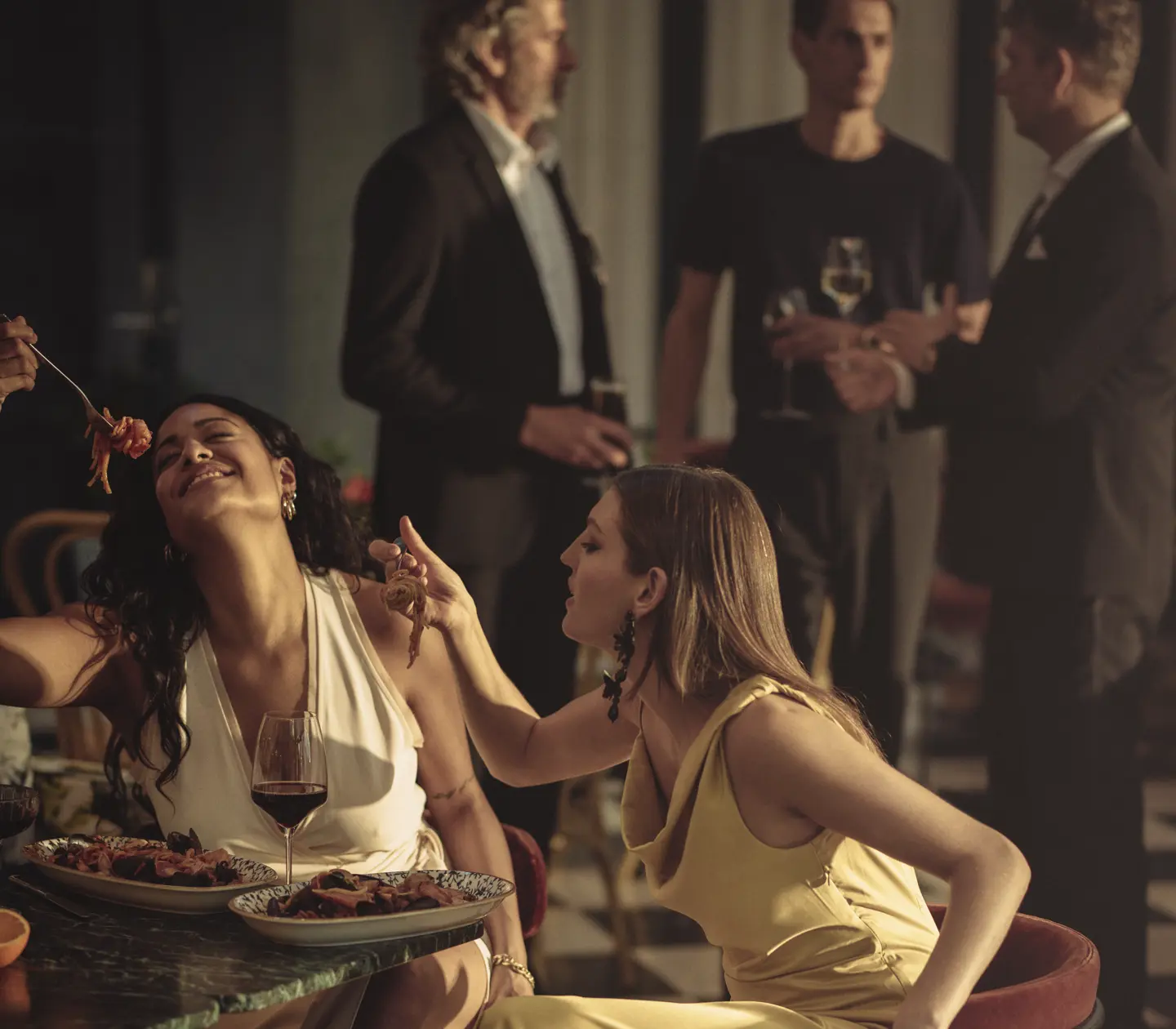 Two women in elegant dresses enjoy a sophisticated dinner at a stylish, upscale restaurant, while three men engage in conversation in the background.