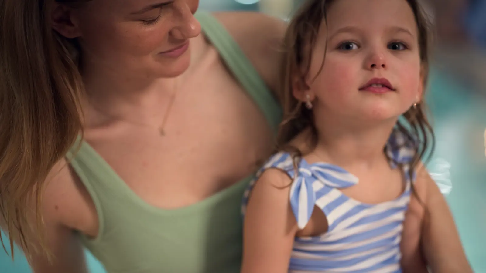 Girl sits on the edge of the pool together with a woman wearing bathing suits