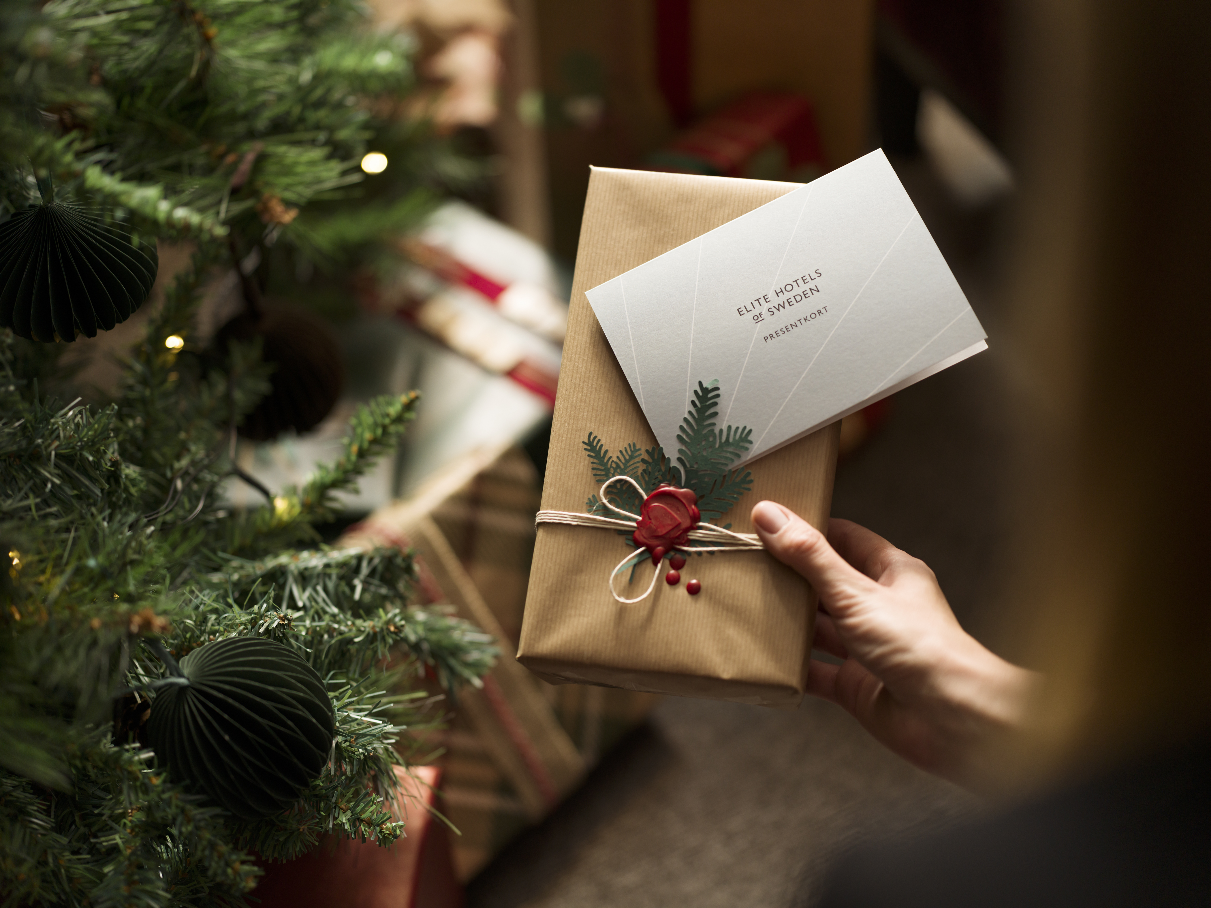 En julklapp med presentkort från Elite Hotels of Sweden och en julgran med julklappar i bakgrunden