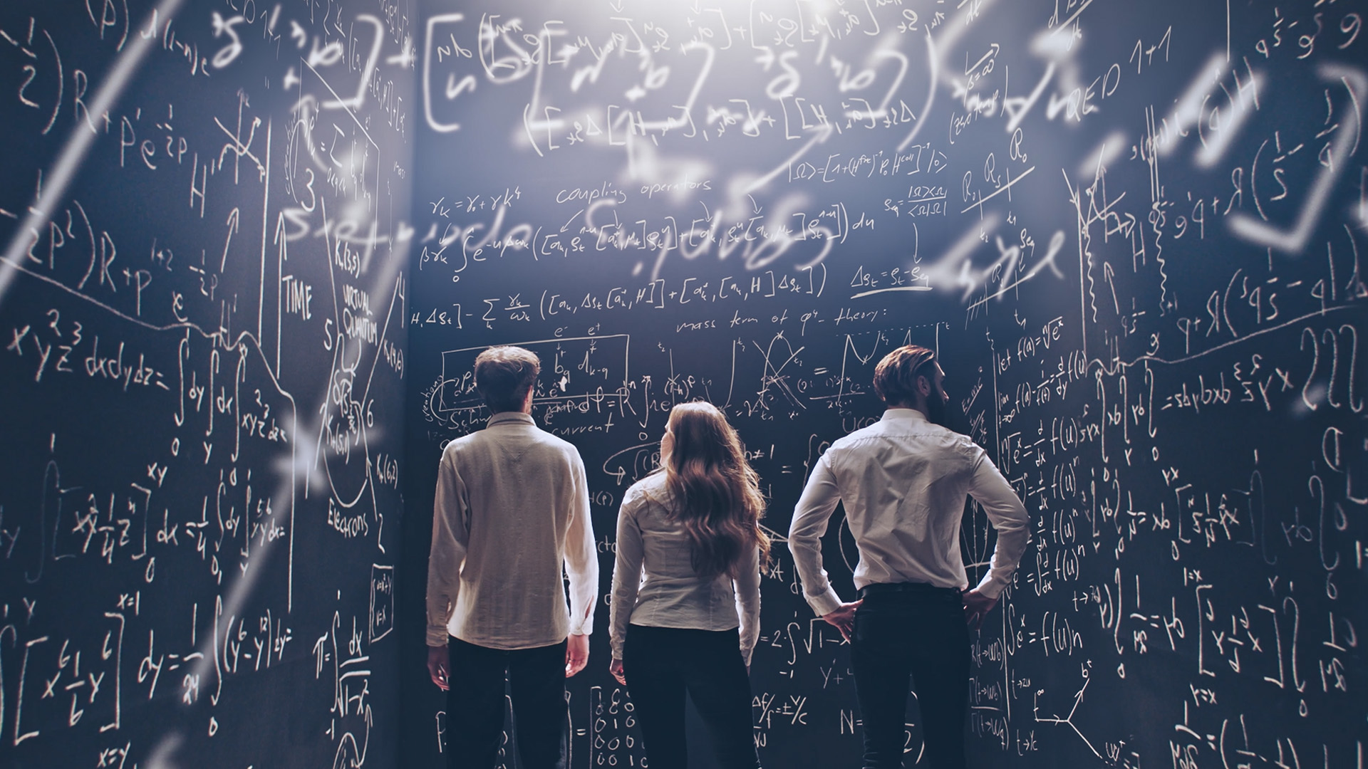 Tre personer i vita skjortor står framför en svart tavla täckt av matematiska formler och ekvationer.