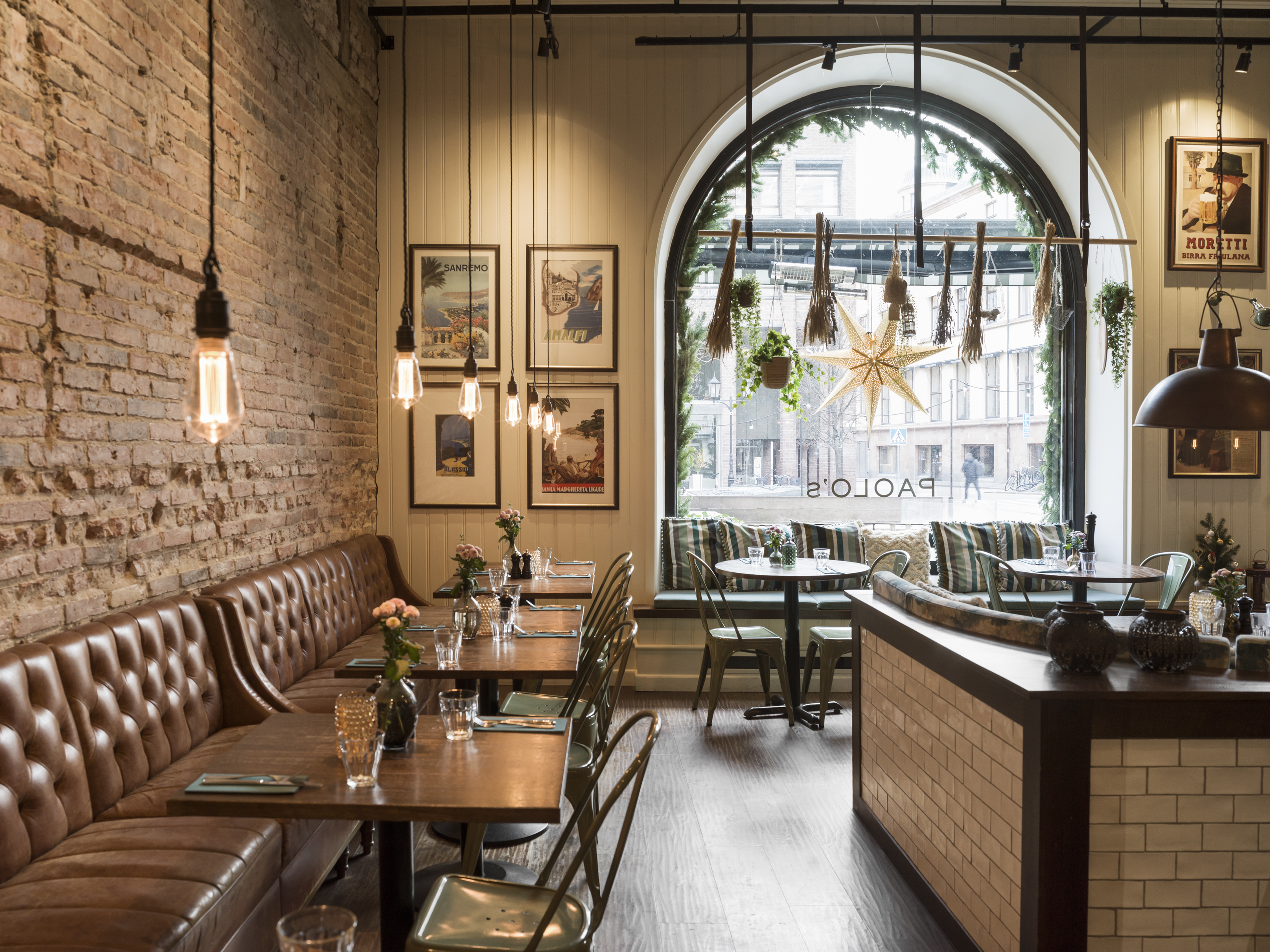 Cozy cafe interior with brick walls, wooden tables, and leather seating. Vintage posters adorn the walls, and large arched windows let in natural light. Hanging lights and indoor plants create a warm, inviting atmosphere.