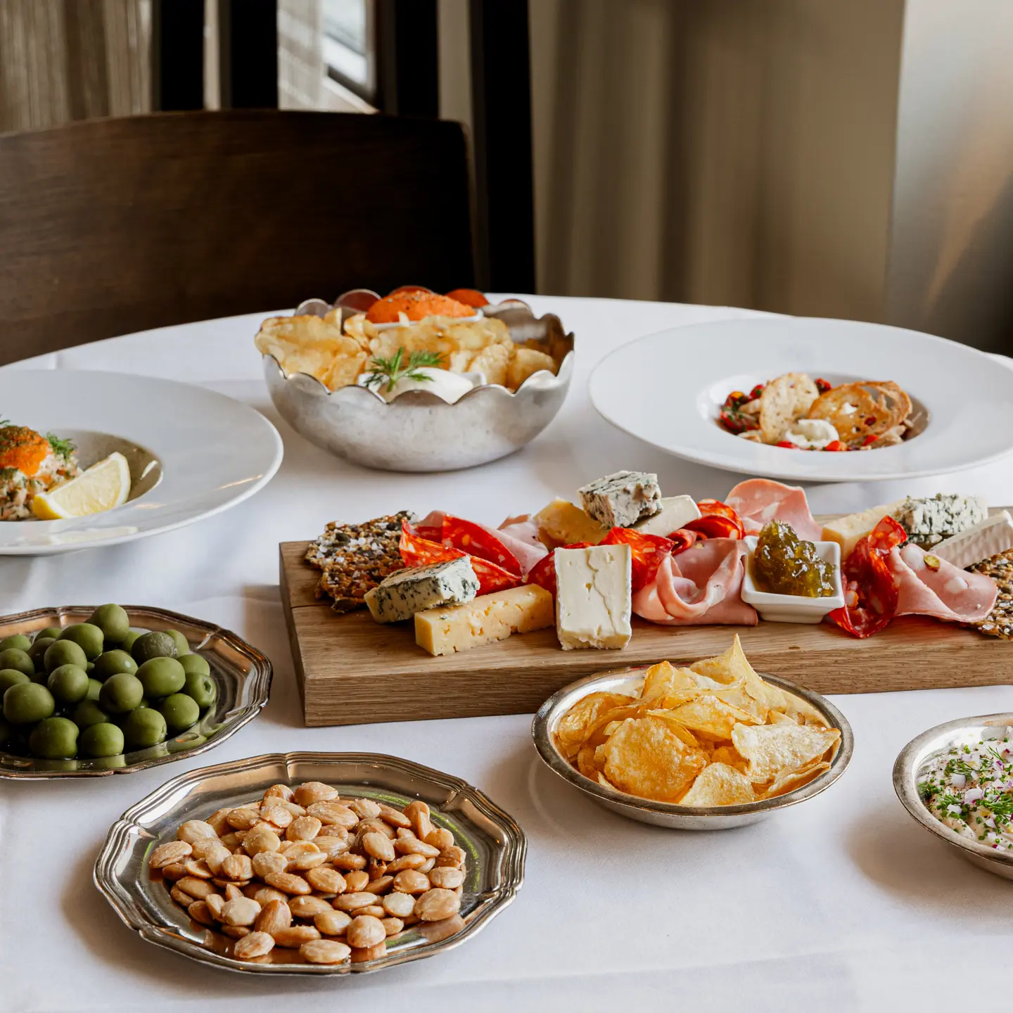 Assorted charcuterie board with cheeses, meats, olives, nuts, chips, and dips on a white tablecloth.