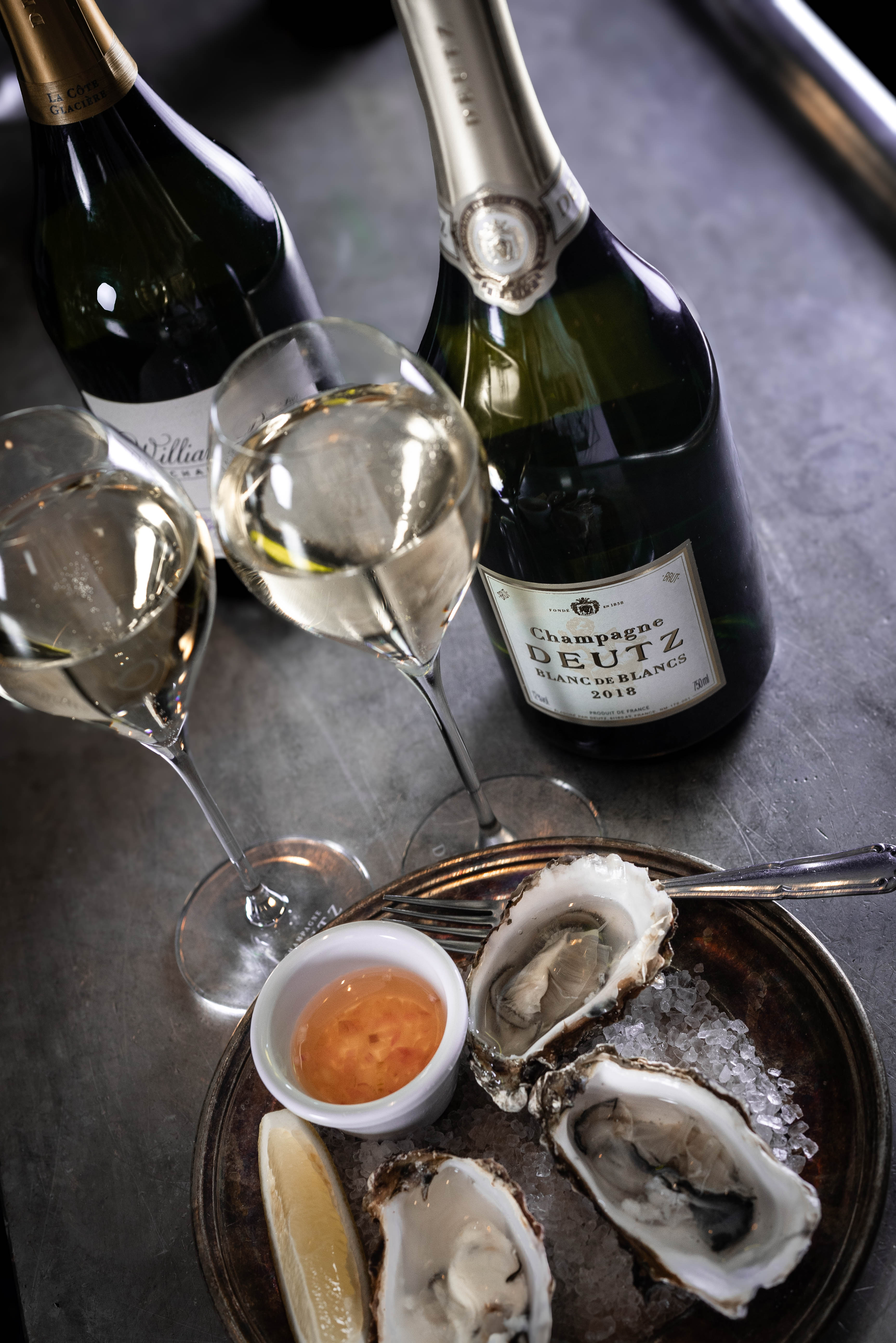 Stilfullt dukat bord på en elegant restaurang, med vinglas och vinflaskor i förgrunden och inredd med klassiska trästolar. Kökspersonal arbetar i bakgrunden.