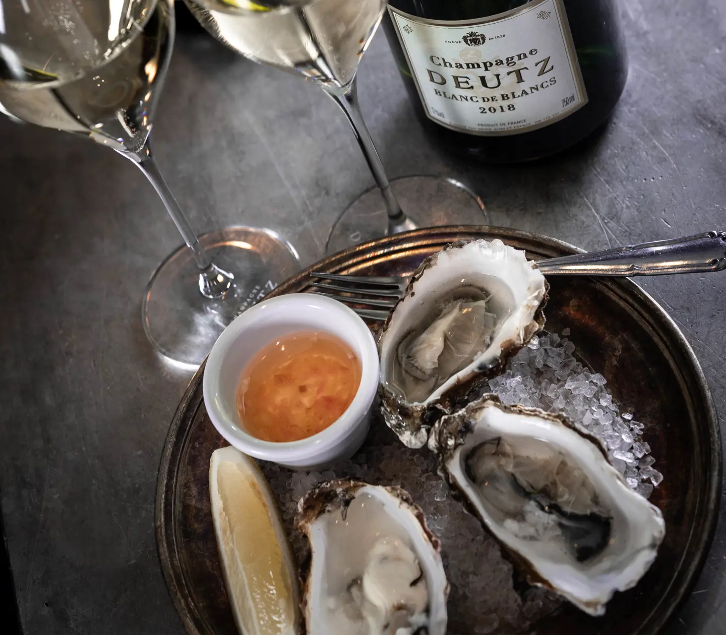 Stilfullt dukat bord på en elegant restaurang, med vinglas och vinflaskor i förgrunden och inredd med klassiska trästolar. Kökspersonal arbetar i bakgrunden.