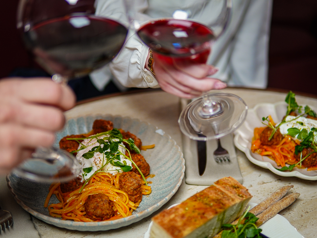 Två personer skålar med vinglas ovanför en tallrik med spaghetti och köttbullar toppade med färska örter, bredvid en brödbit och ost på ett bord.