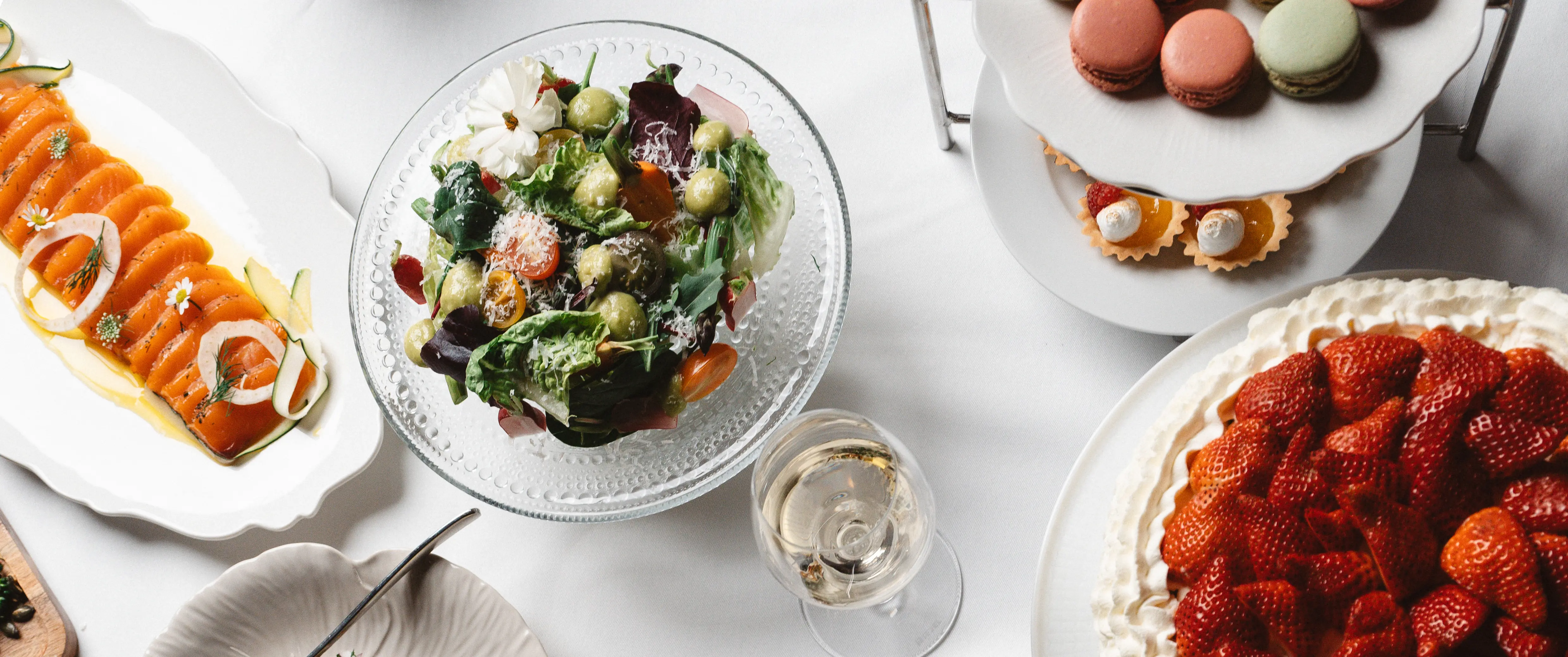 Lyxig buffé med rökt lax, sallad, macarons och jordgubbstårta serverad på ett elegant bord med ett glas vitt vin.