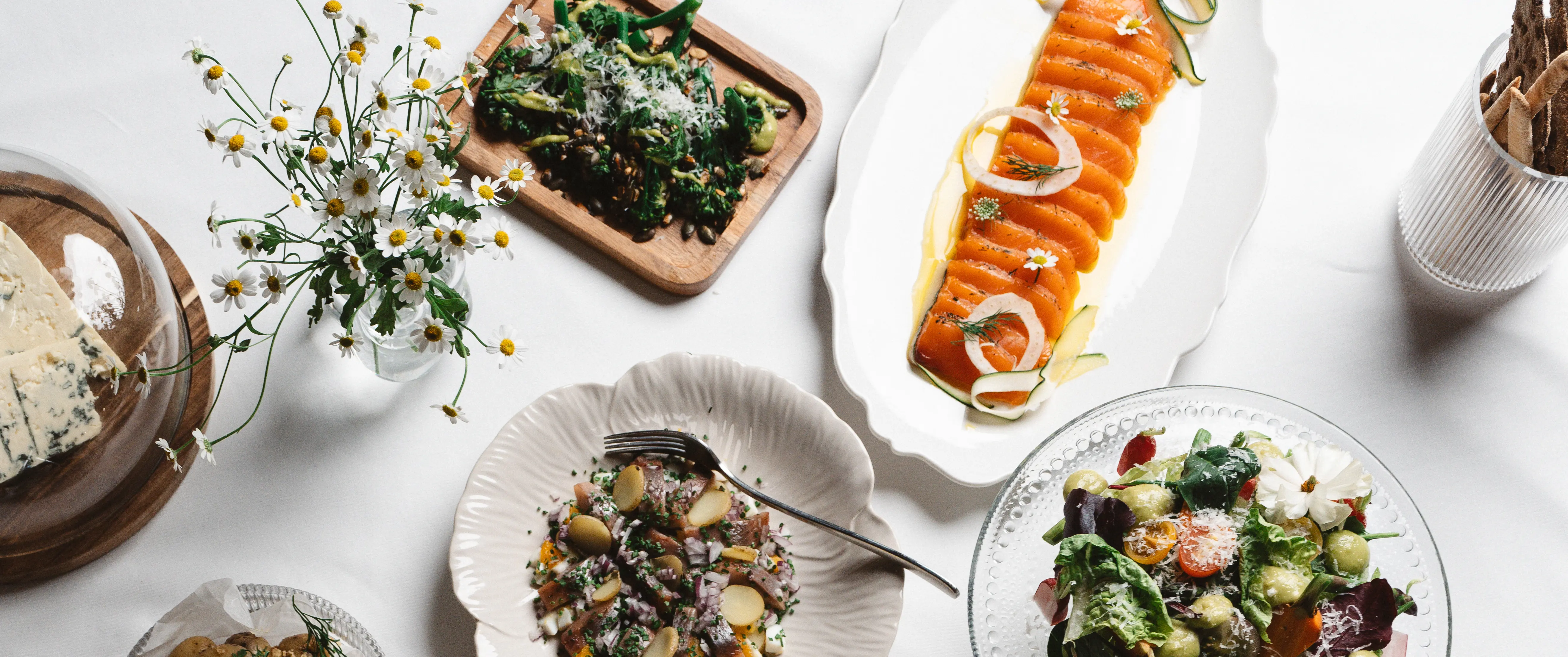 Svenskt smörgåsbord med rökta laxskivor, färsk sallad, ostar, bröd och grönsaksrätt på ett snyggt dukat bord med blommor.