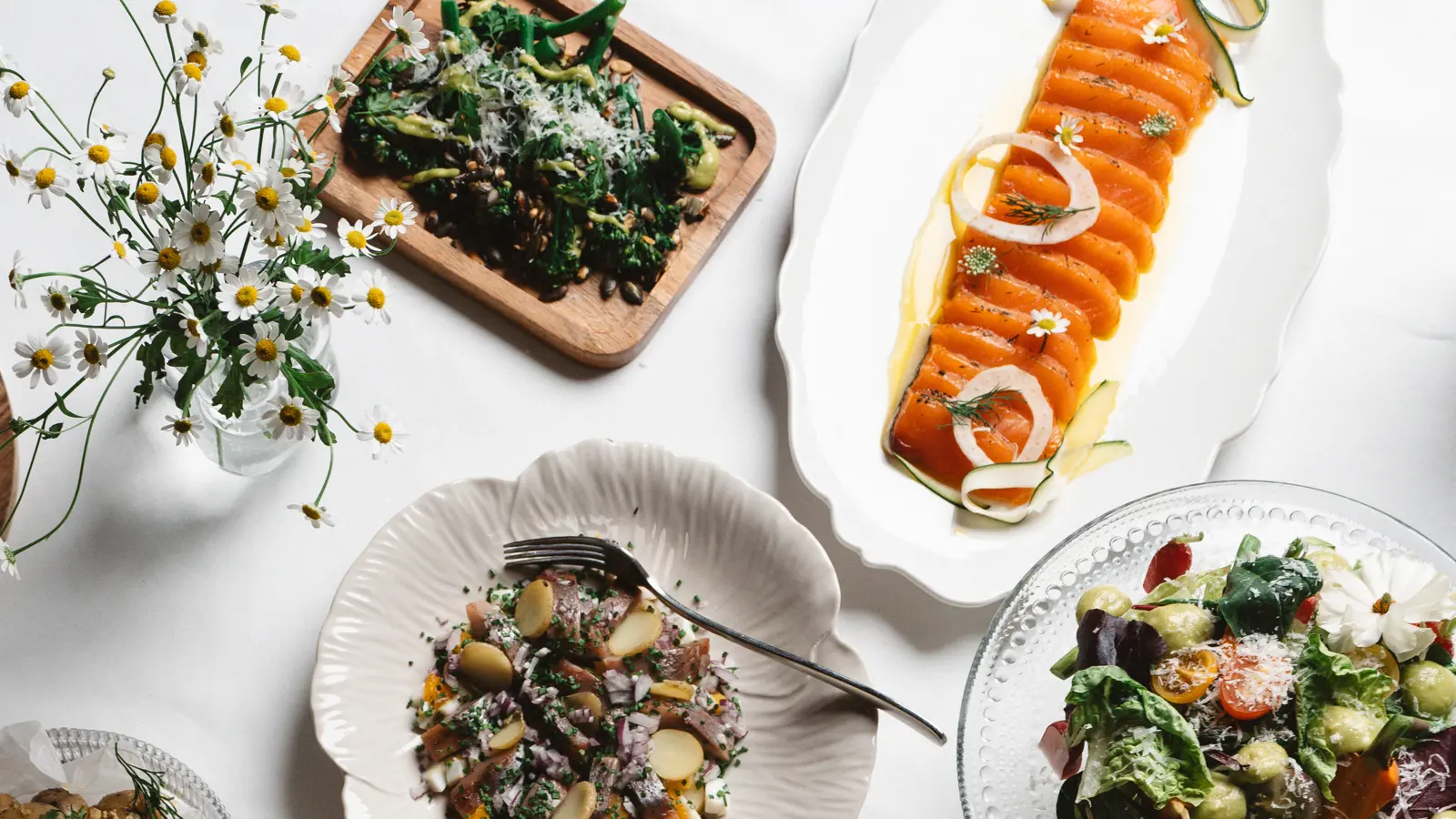 Svenskt smörgåsbord med rökta laxskivor, färsk sallad, ostar, bröd och grönsaksrätt på ett snyggt dukat bord med blommor.