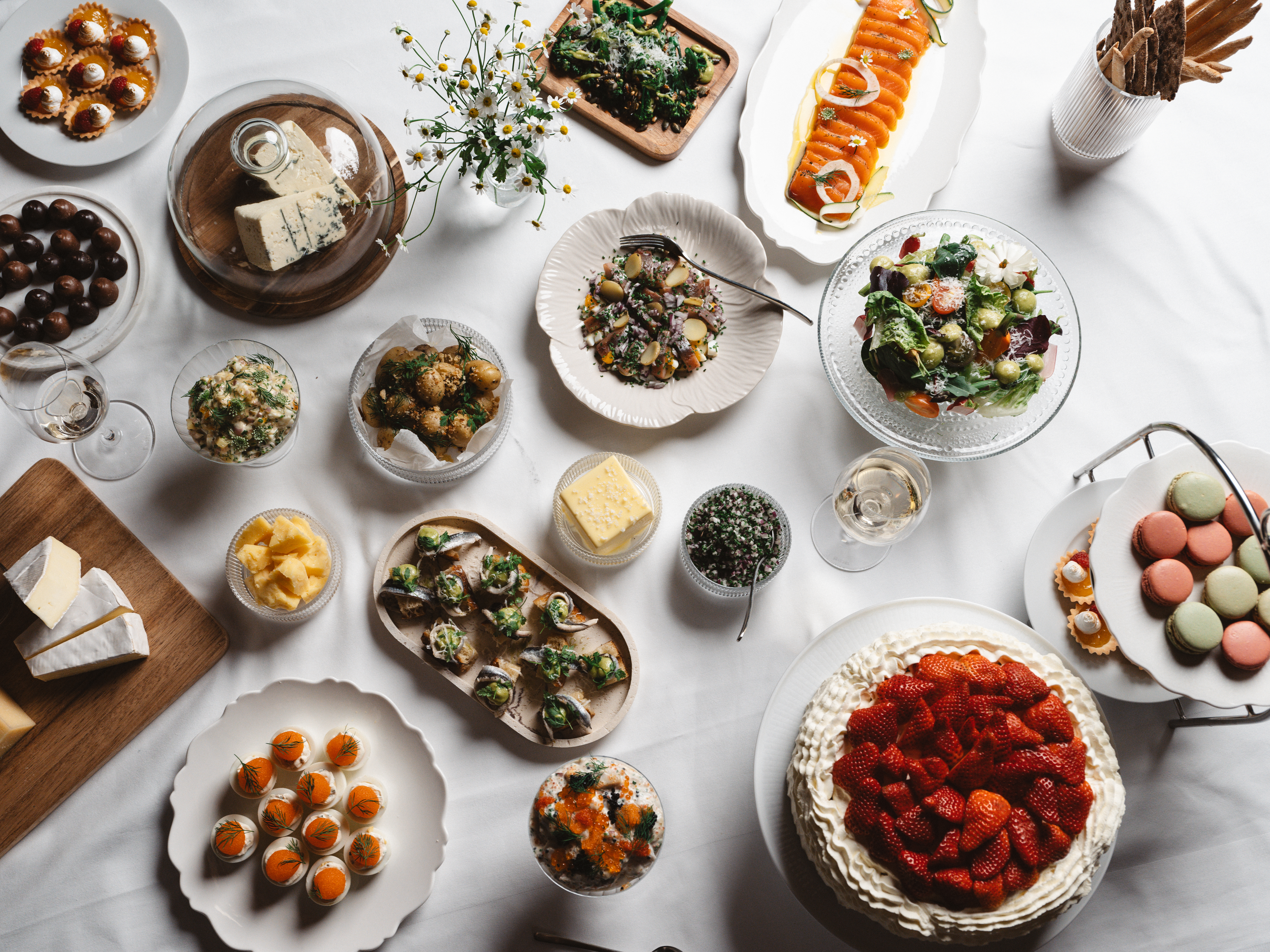 Svenskt smörgåsbord med olika delikatesser inklusive inlagd lax, sallad, bröd, ostar, tårta med jordgubbar, och macarons på ett uppdukat bord.