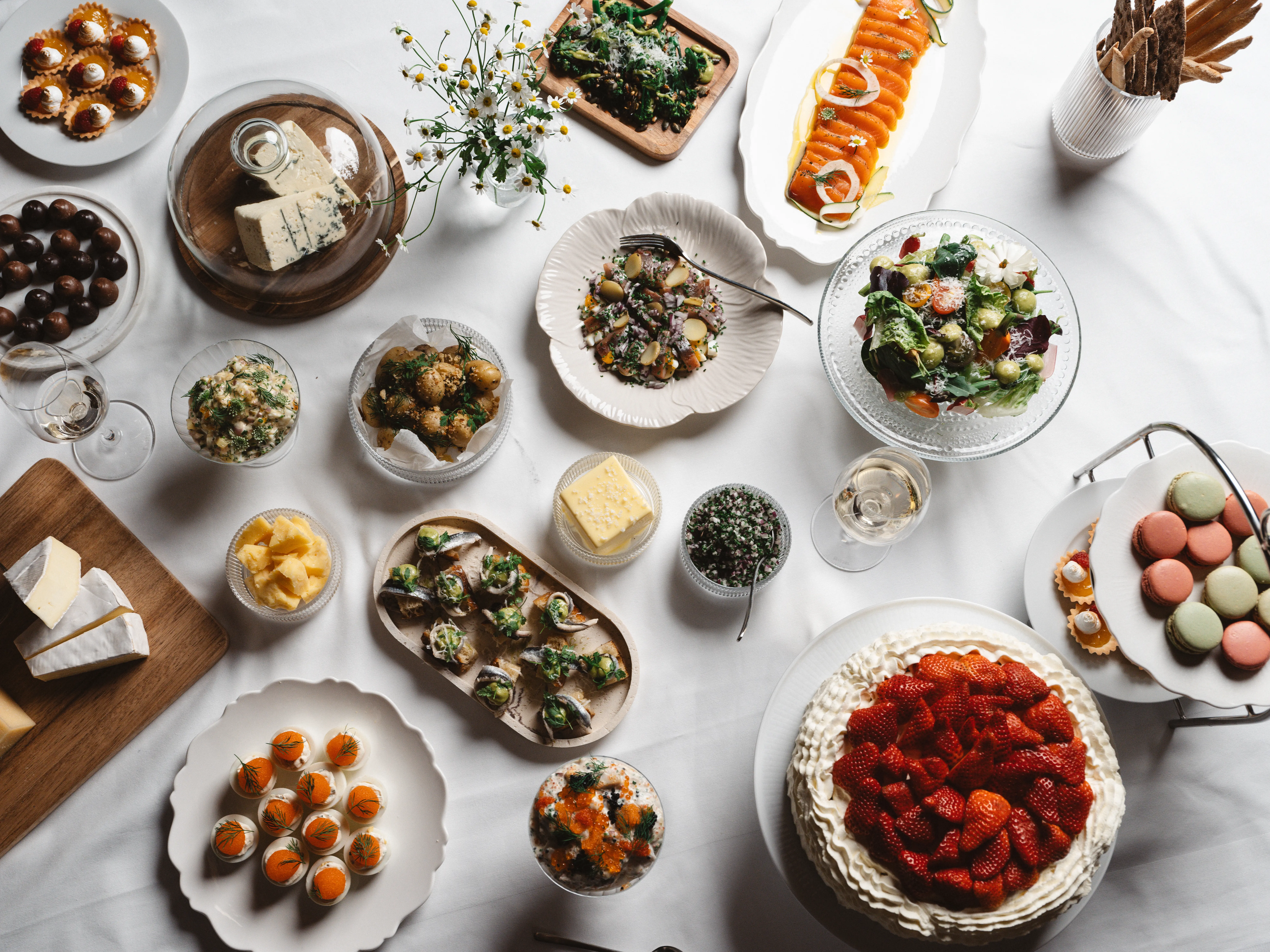 Svenskt smörgåsbord med olika delikatesser inklusive inlagd lax, sallad, bröd, ostar, tårta med jordgubbar, och macarons på ett uppdukat bord.