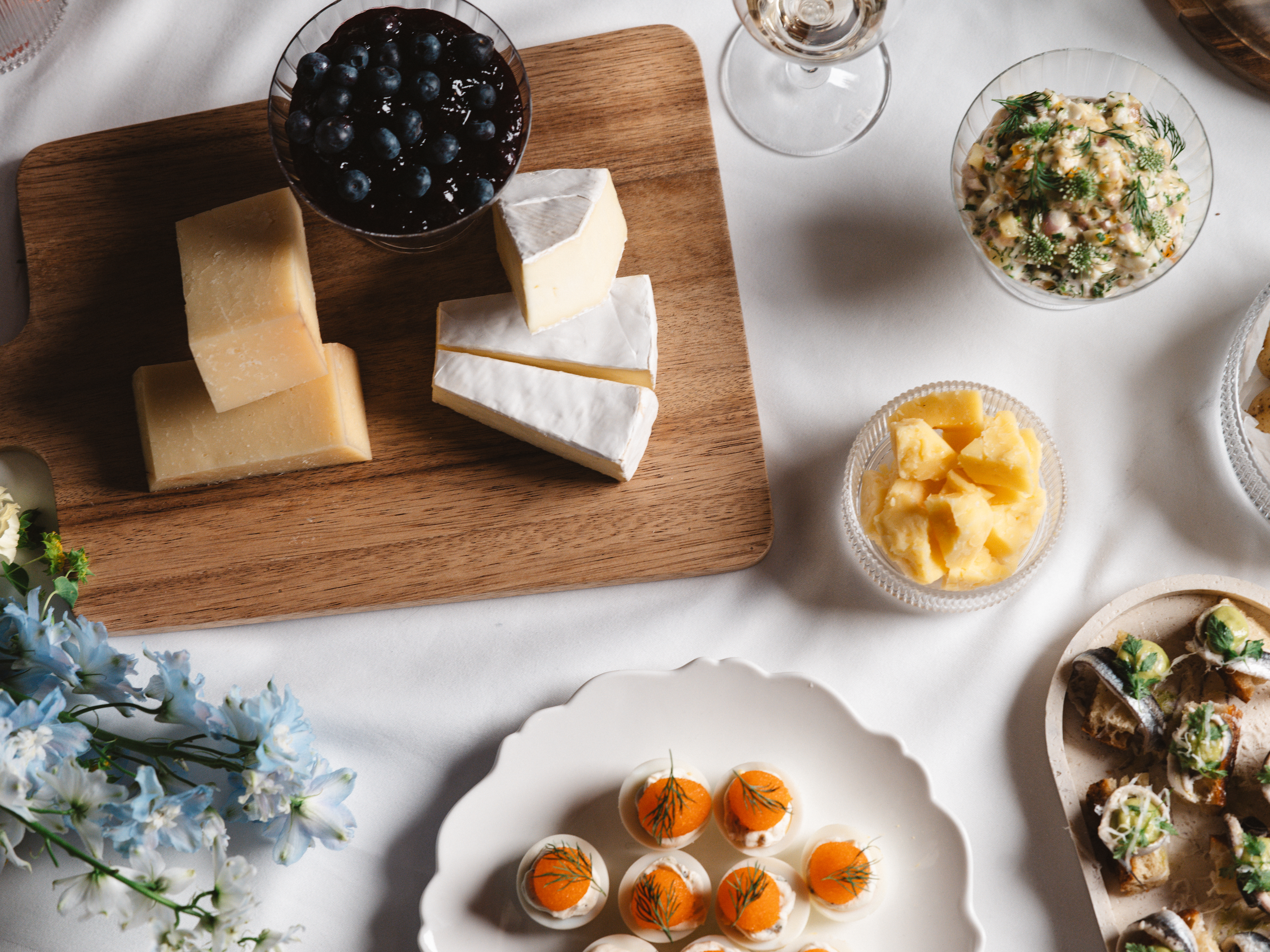 Cheese platter with blue cheese under a glass dome on a wooden platter, surrounded by chocolate pralines, small pastries and a vase of flowers on a set table.