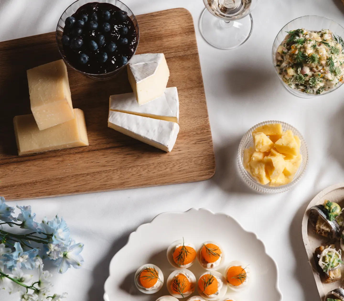 Cheese platter with blue cheese under a glass dome on a wooden platter, surrounded by chocolate pralines, small pastries and a vase of flowers on a set table.