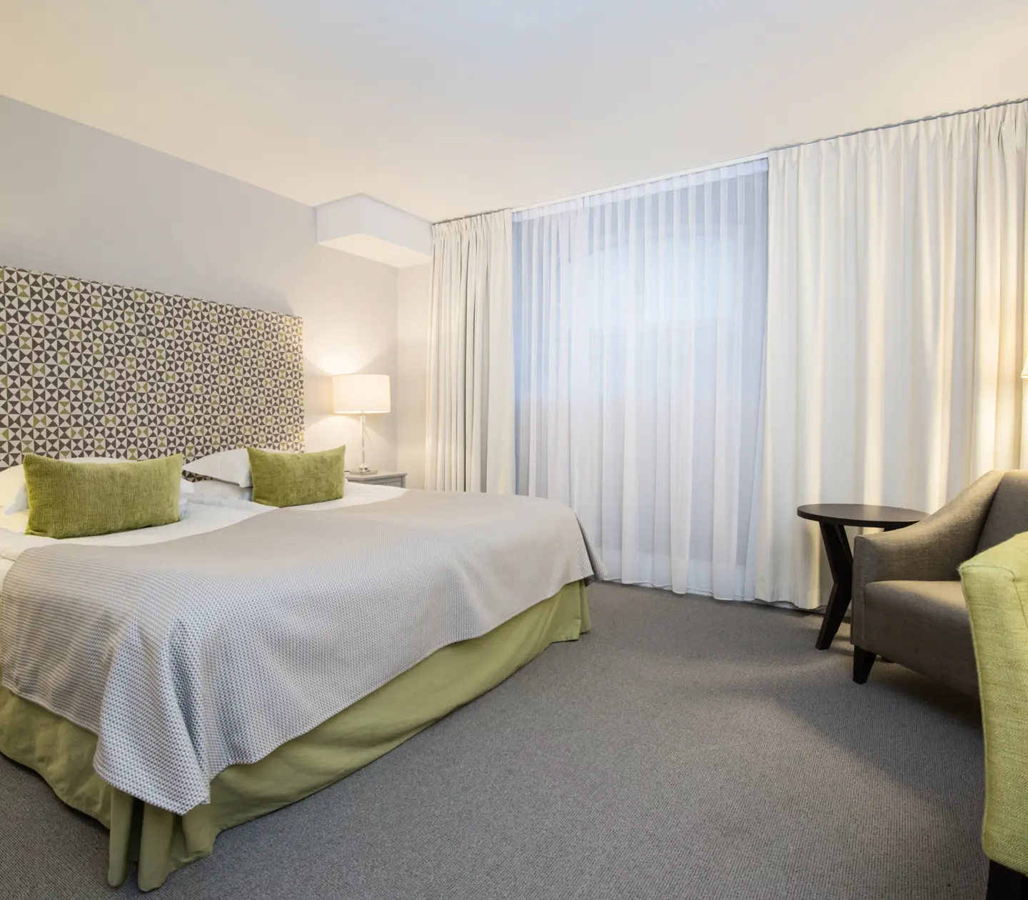 Ljust hotellrum med säng, fåtölj och gardier
