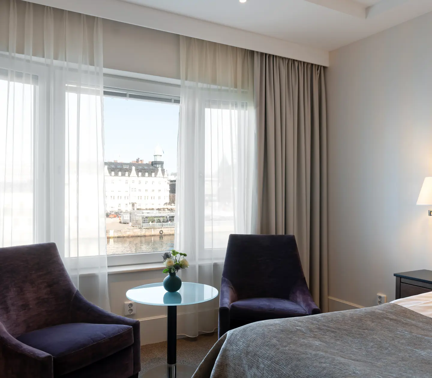 Ljust hotellrum med fåtöljer och fönster med utsikt över hamnen