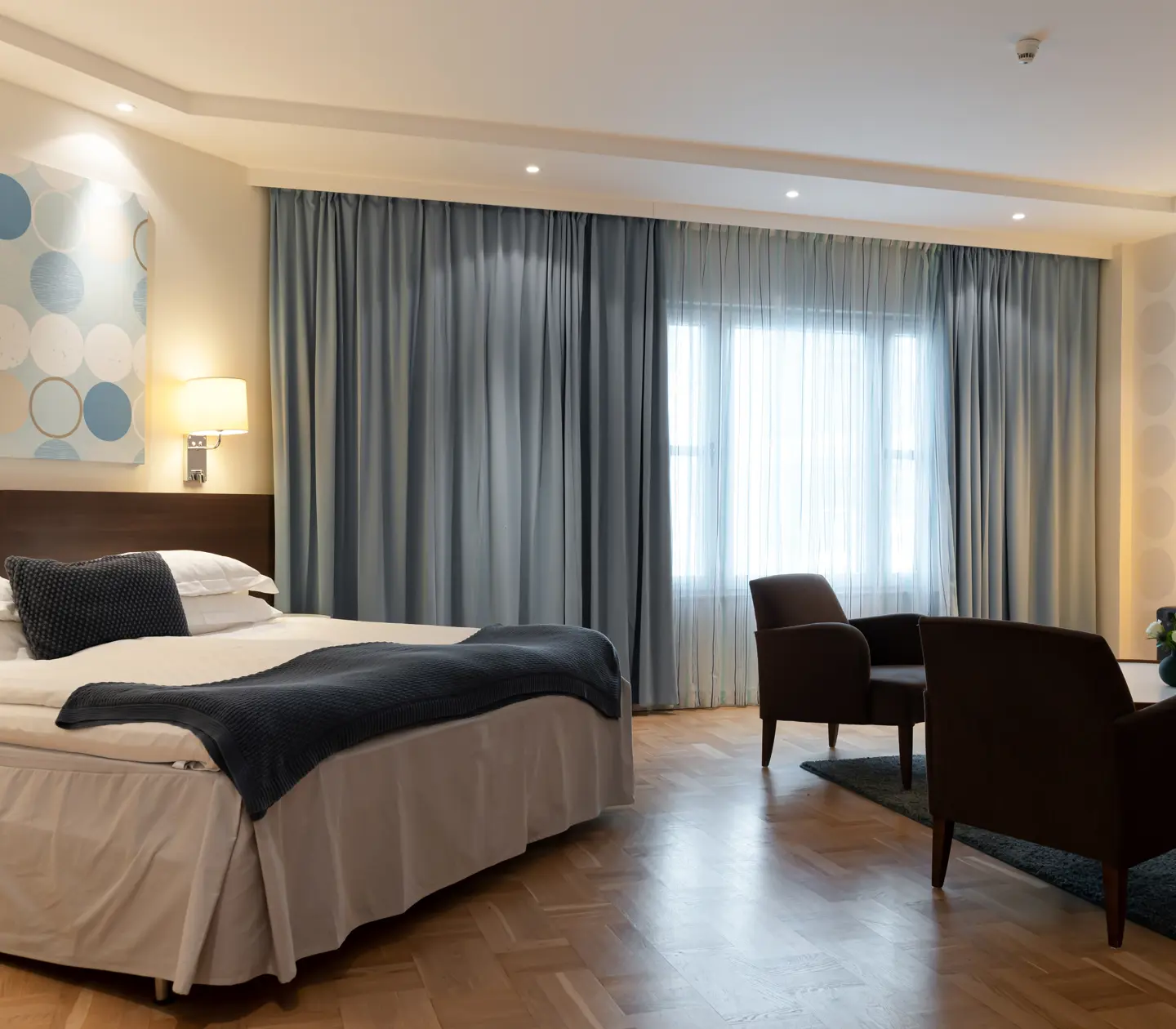 Ljust hotellrum med dubbelsäng, soffgrupp och blå gardiner
