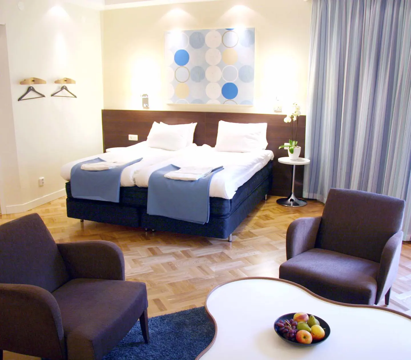 Ljust hotellrum med säng, fåtöljer och blå gardiner