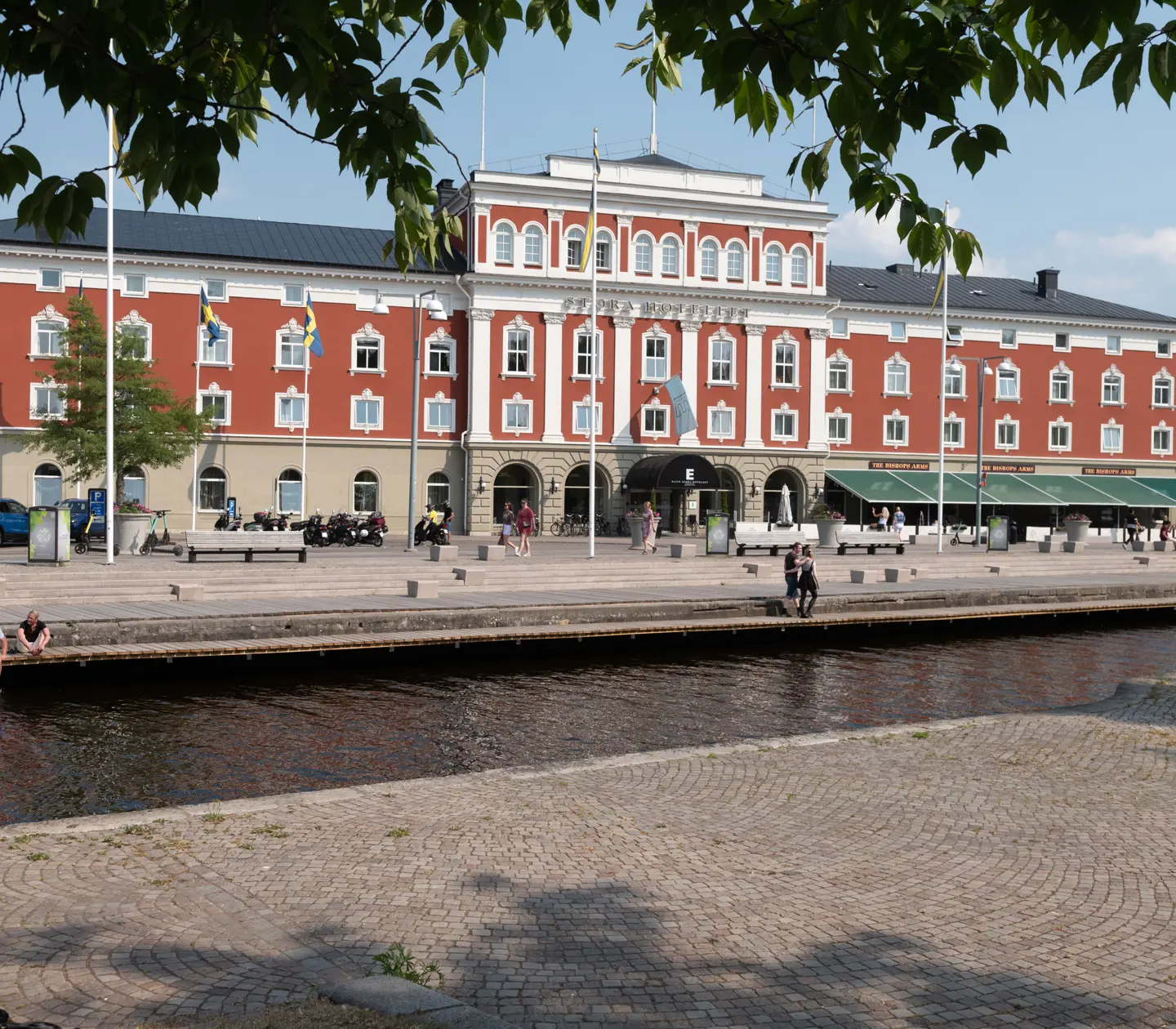 Rött historiskt hotell vid en kanal i centrala Jönköping, Sverige, med folk som promenerar och sitter vid vattnet under en klar himmel