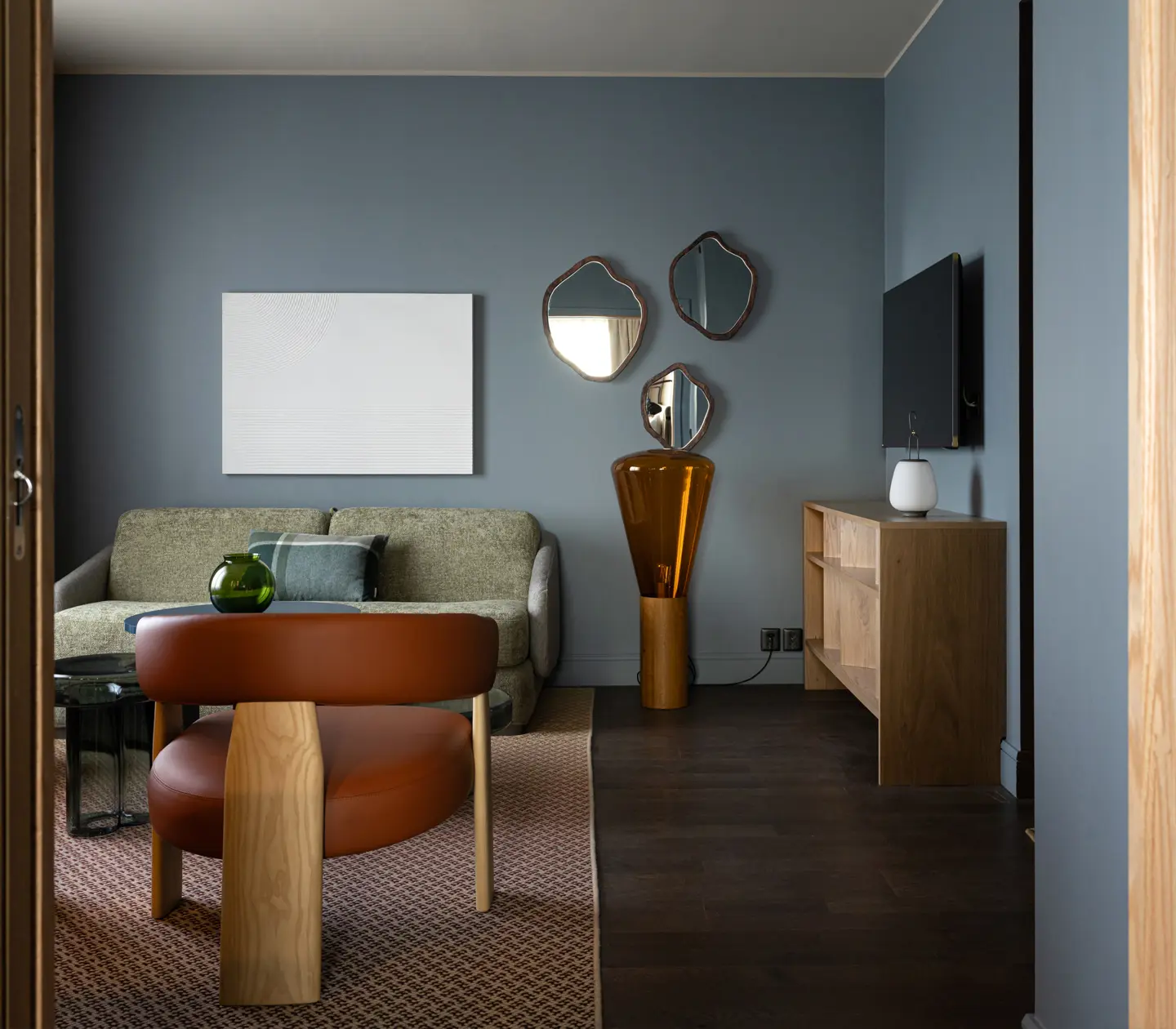 Vardagsrum med modern inredning, grå väggar och trämöbler. En grön soffa, läderfåtölj och dekorativa speglar skapar en stilfull atmosfär. En stor tv och vas på hylla för komplett look