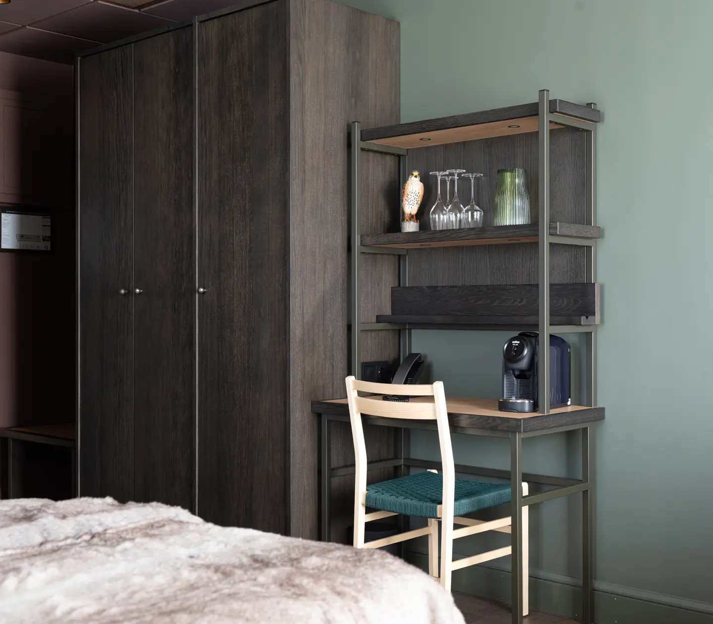 Hotellrum med mörkt träskåp, hylla med glas och kaffemaskin, samt arbetsyta med stol mot grön vägg