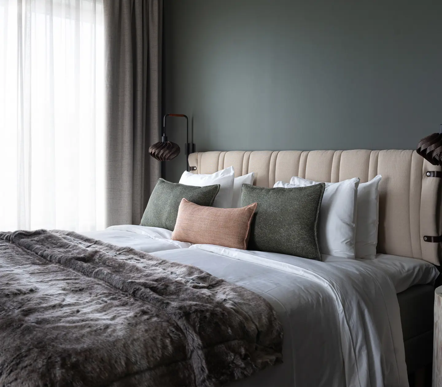 Elegant sovrum med modern inredning. En bekväm säng med beige klädsel, flera dekorativa kuddar och ett grått överkast. Mjukt naturligt ljus filtreras genom långa gardiner